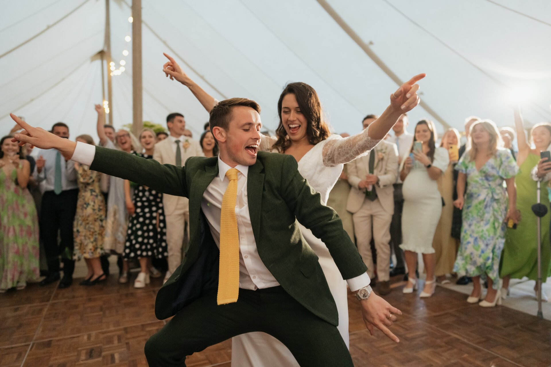 A couple in fancy clothes is dancing with lots of energy at a wedding party, while guests cheer them on under a tent.