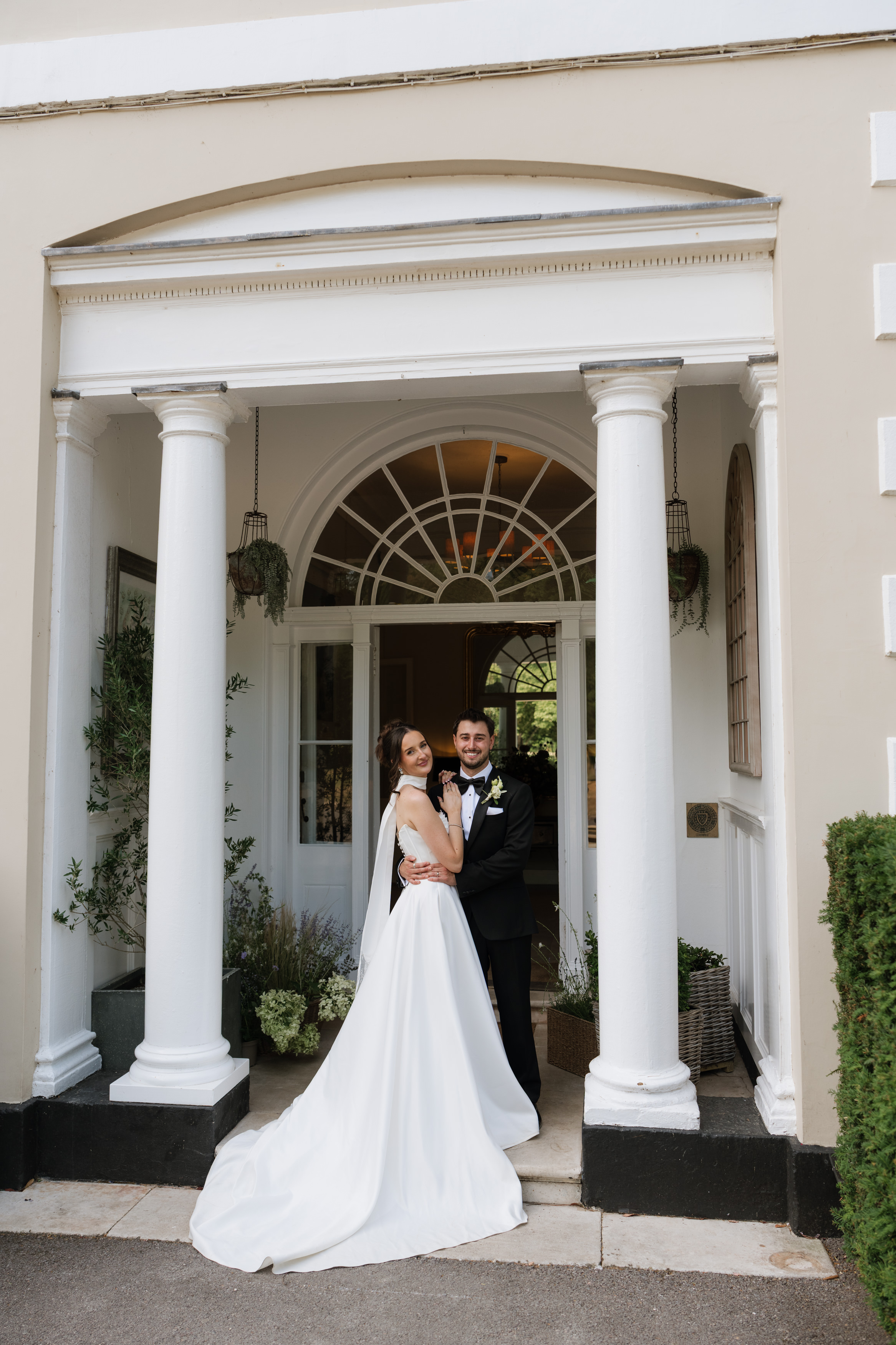 A bride and groom smile as they stand together under a white arch with columns in front of a building entrance.