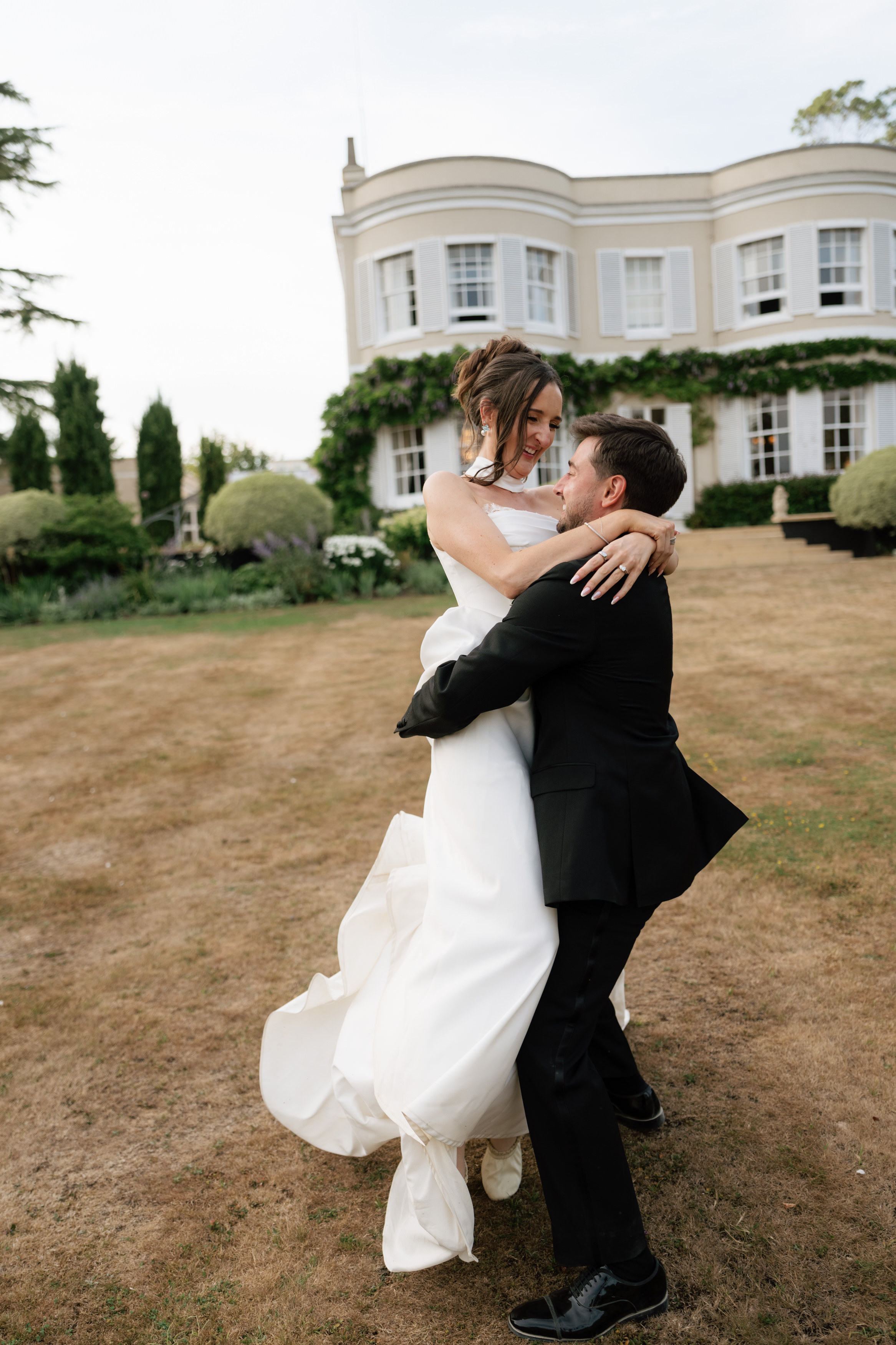 A groom wearing a black suit lifts and hugs his bride, who’s in a white dress, as they stand on the grass in front of a large white house.