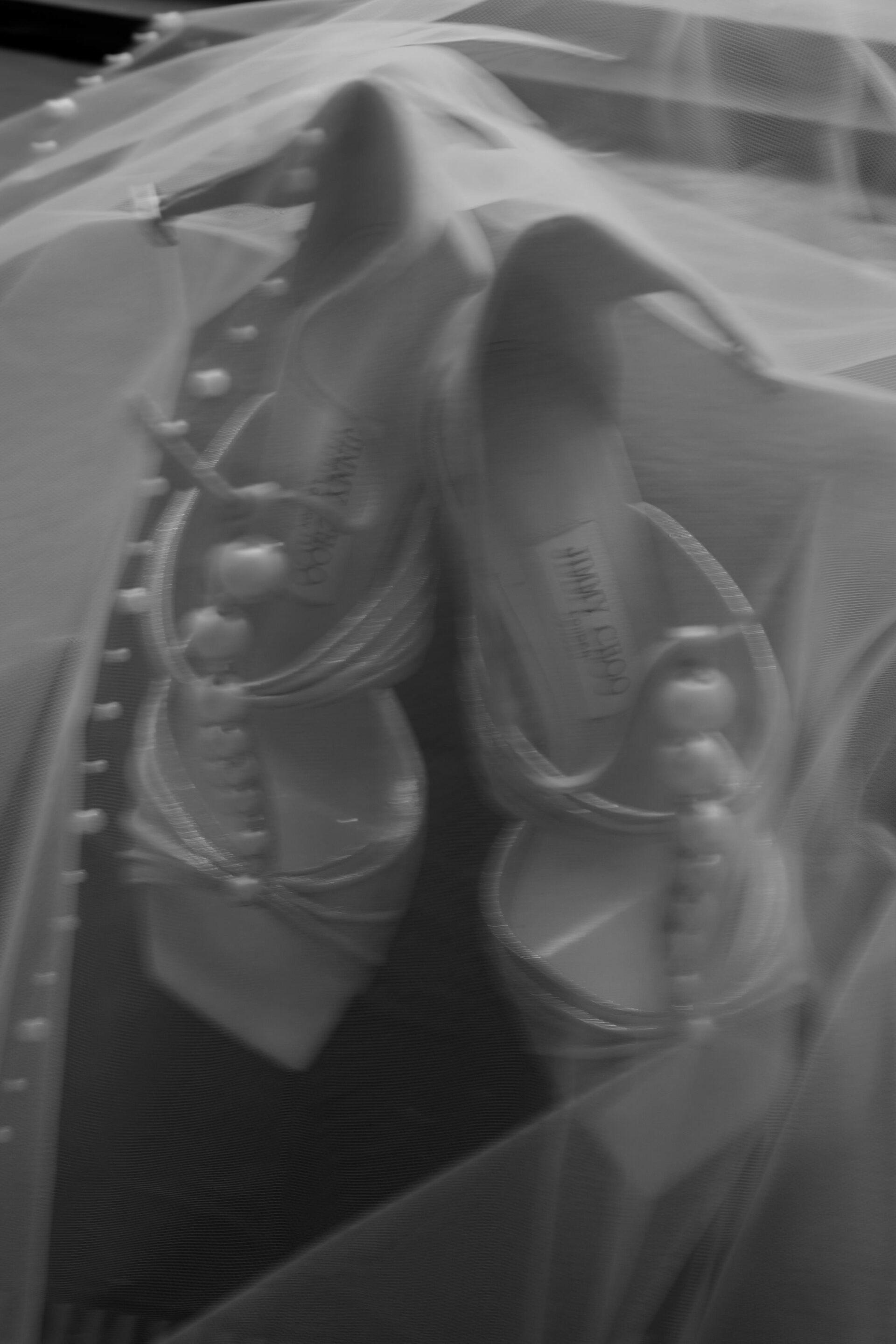 A black and white photo showing high-heeled shoes decorated with pearls, partly covered by a see-through fabric.