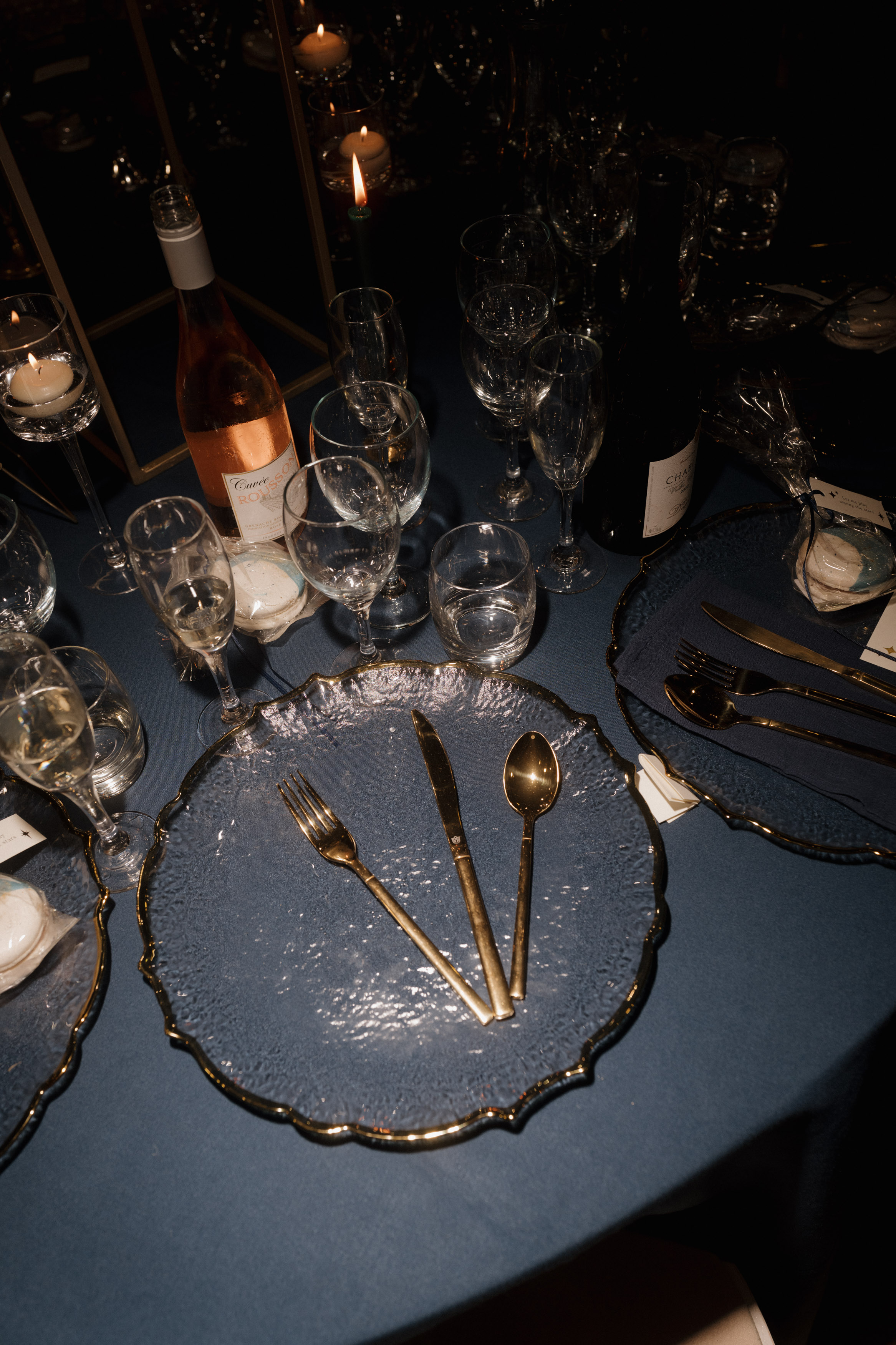 A dining table is set up with gold-colored utensils and a clear glass plate. There are wine glasses, a bottle of wine, and lit candles on a blue tablecloth.