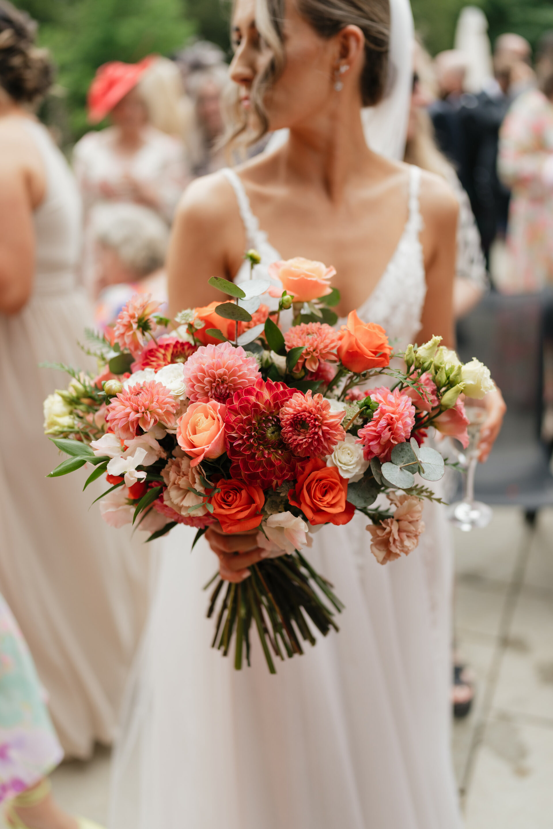 A bride in a white dress holds a colorful bunch of orange, pink, and white flowers at an outdoor event.