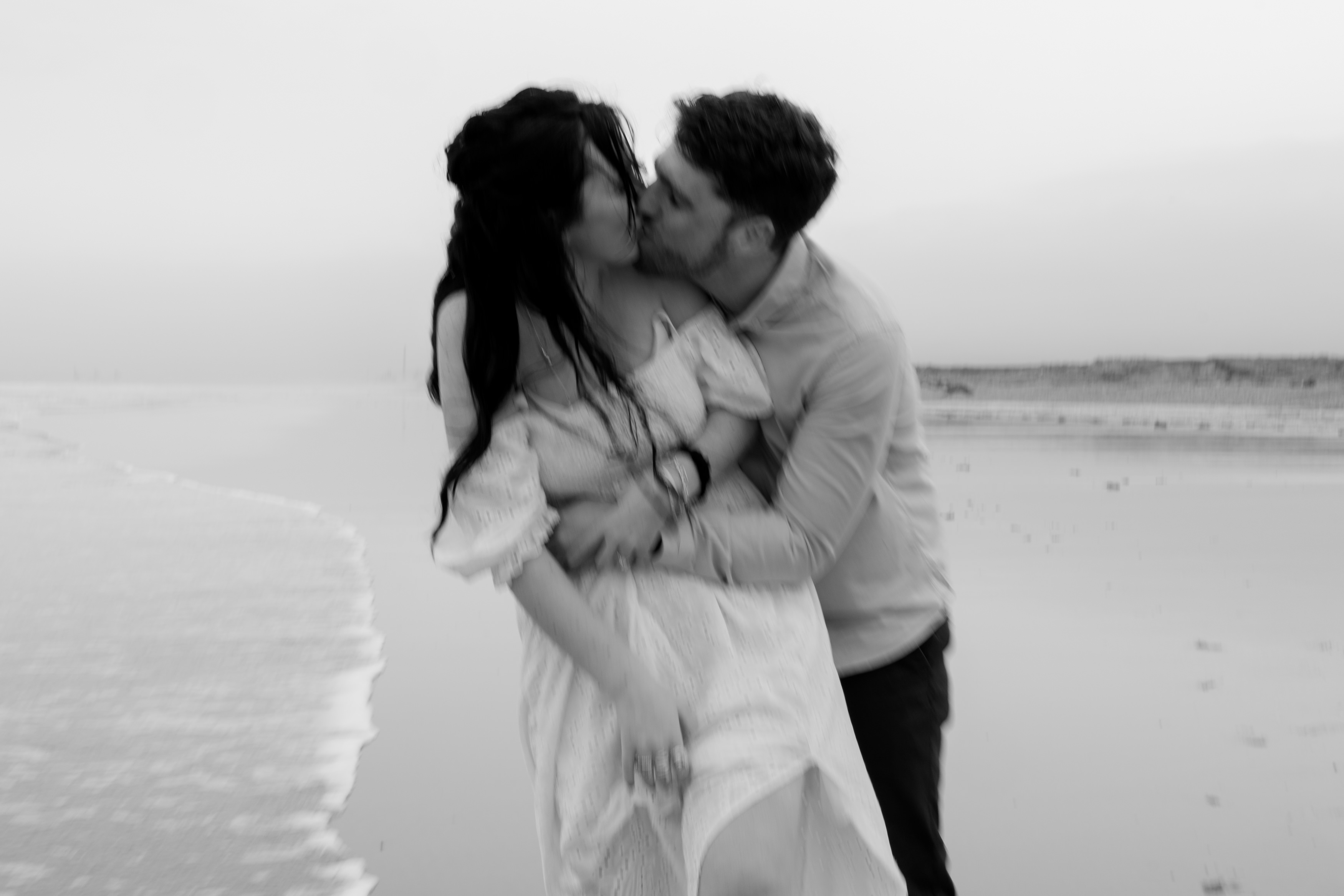 A couple is kissing on an empty beach, with soft waves behind them. The picture is in black and white.