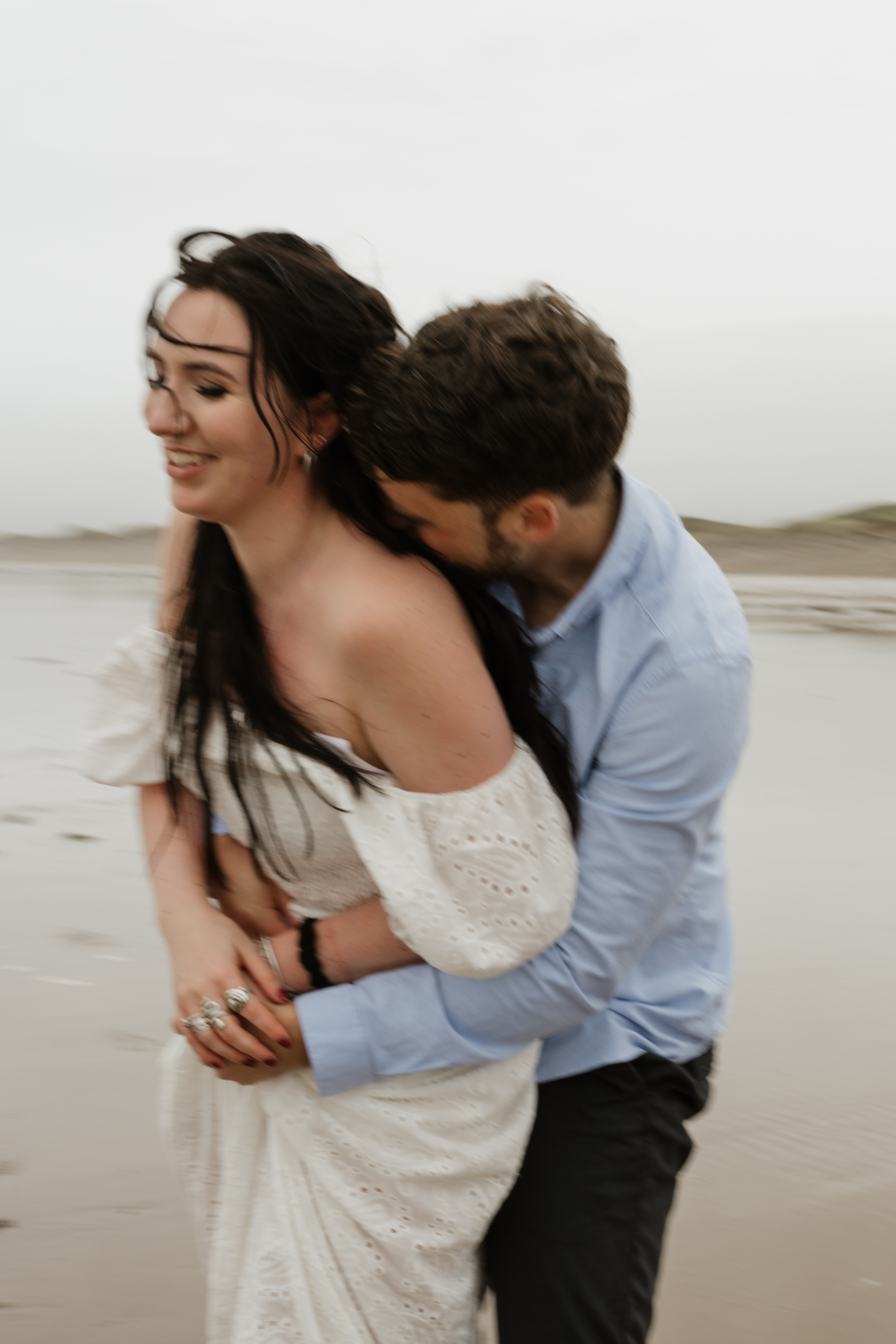 A couple is having fun on a windy beach. The guy hugs the girl from behind while she laughs. She's wearing a white dress, and he's in a light blue shirt. The sky is cloudy.