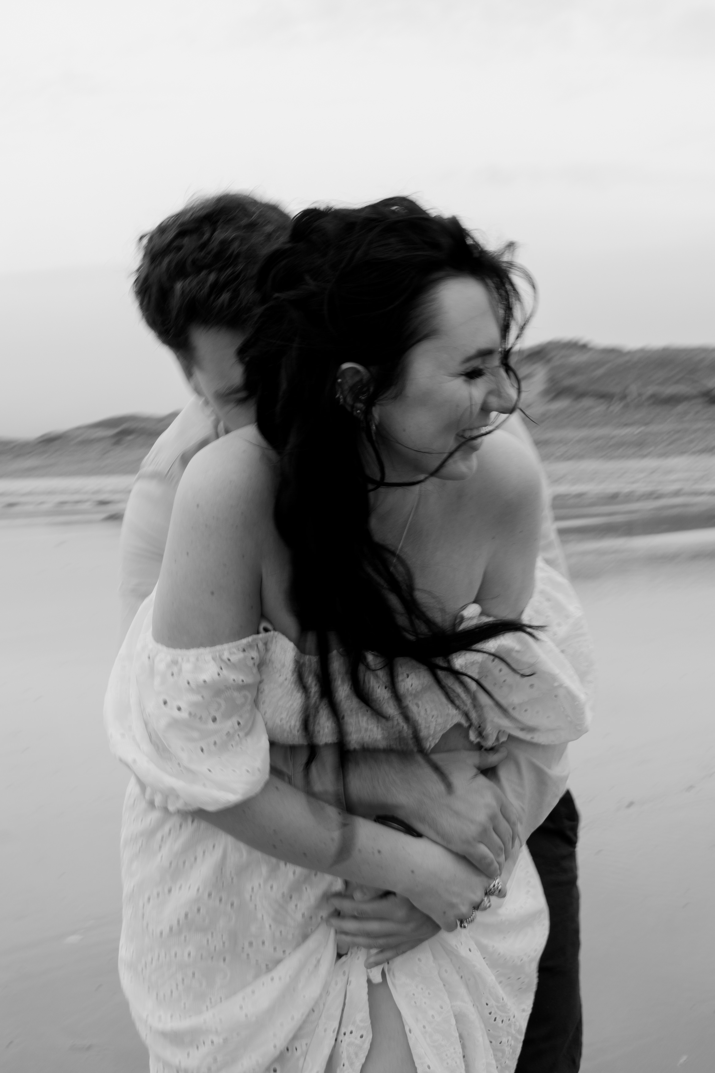 In the black-and-white photo, there's a couple at the beach. The man is hugging the woman from behind, and they're both laughing. She's wearing a dress that sits off her shoulders. In the background, you can see sand dunes and a cloudy sky.