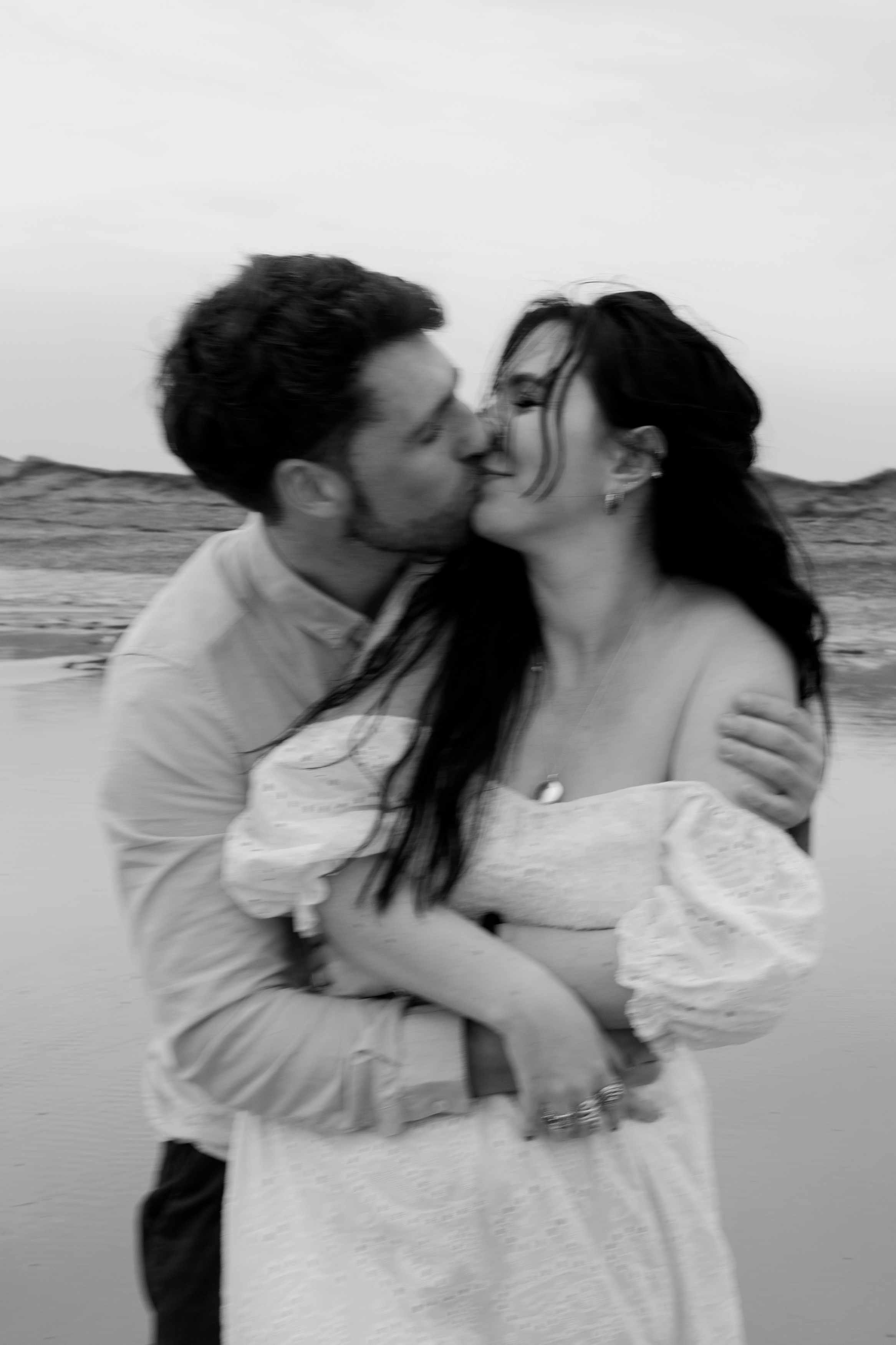A couple is hugging and kissing on the beach, with the man softly holding the woman from behind. The scene feels laid-back and intimate.