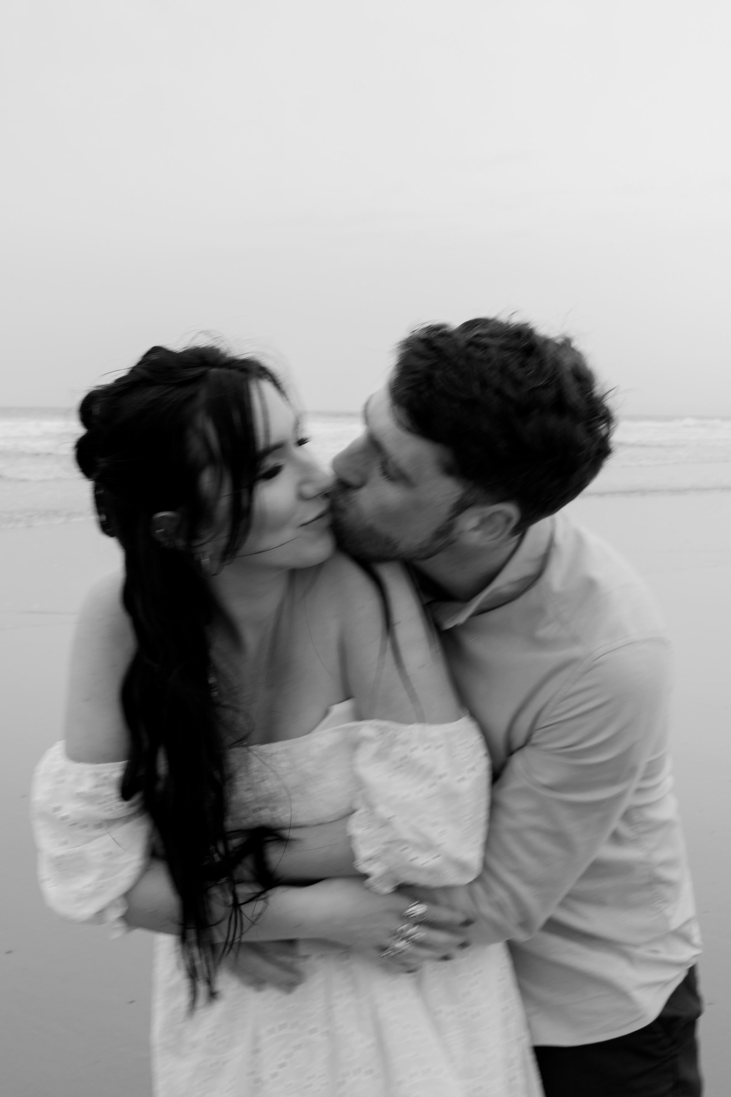 A couple is kissing on the beach with the ocean behind them, and the photo is in black and white.