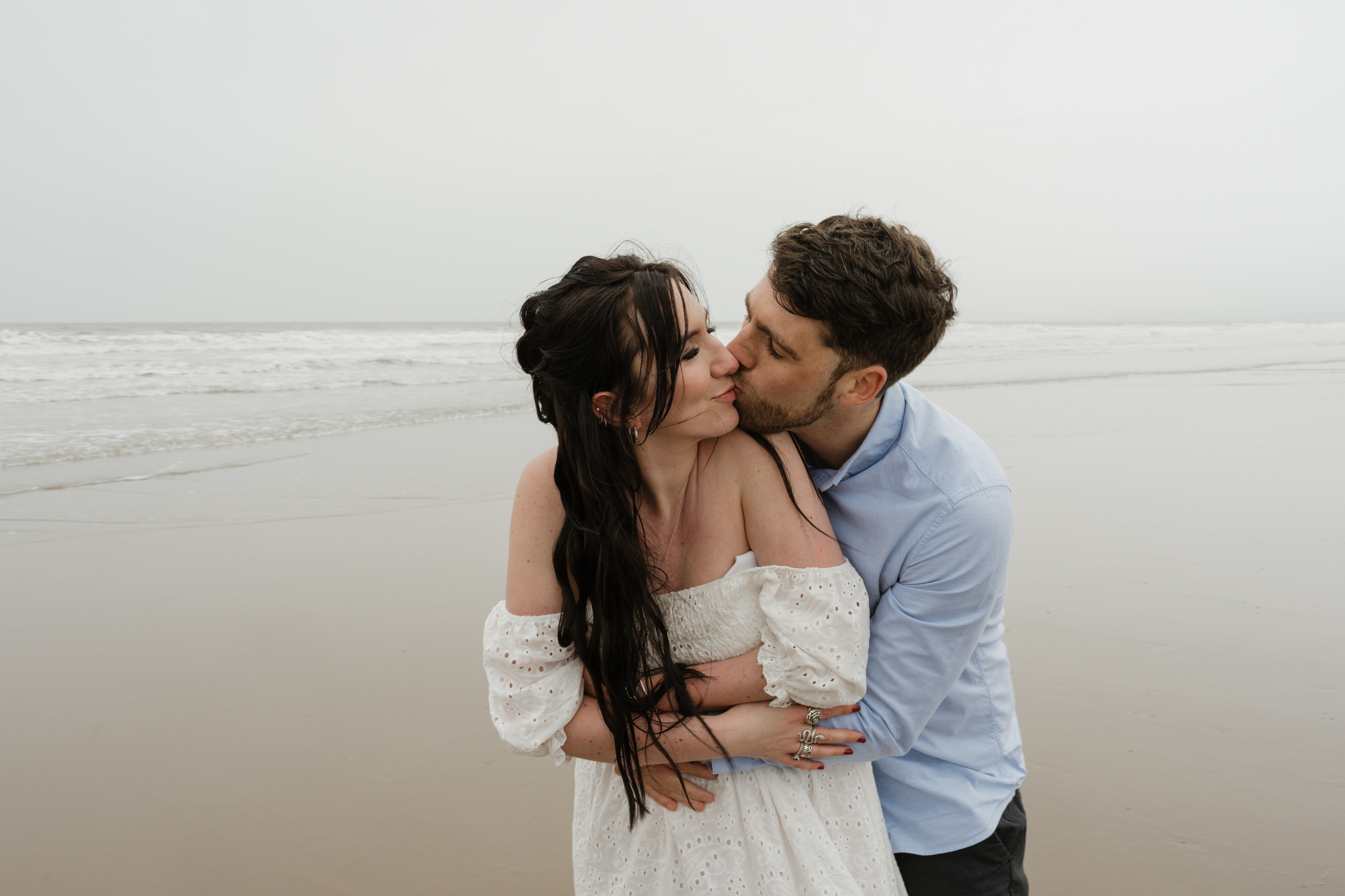A couple is hugging and kissing on a beach. The woman is in a white dress, and the man has on a light blue shirt. You can see the ocean waves behind them.