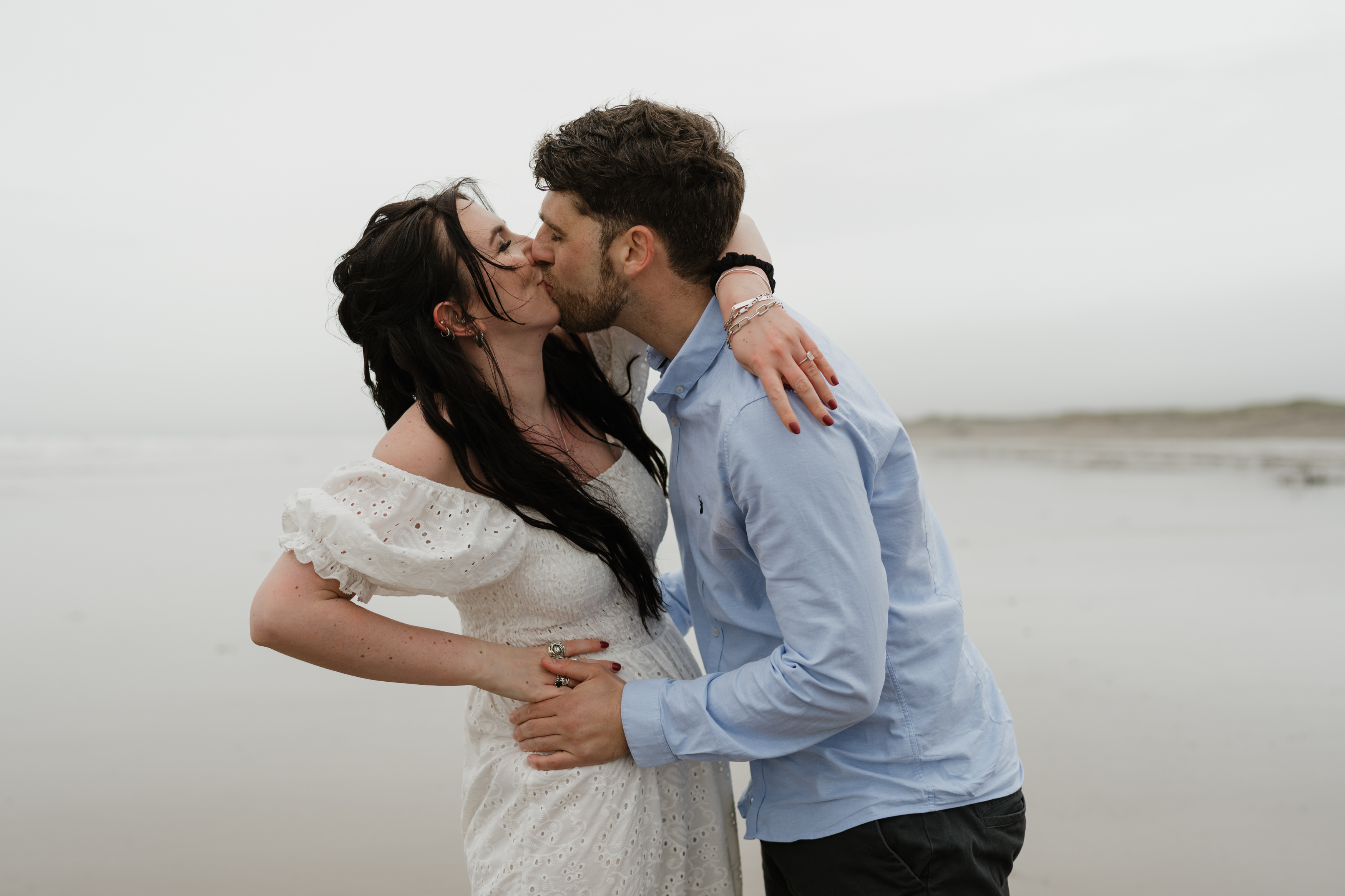 A woman in a white dress and a man in a light blue shirt are kissing on the beach.