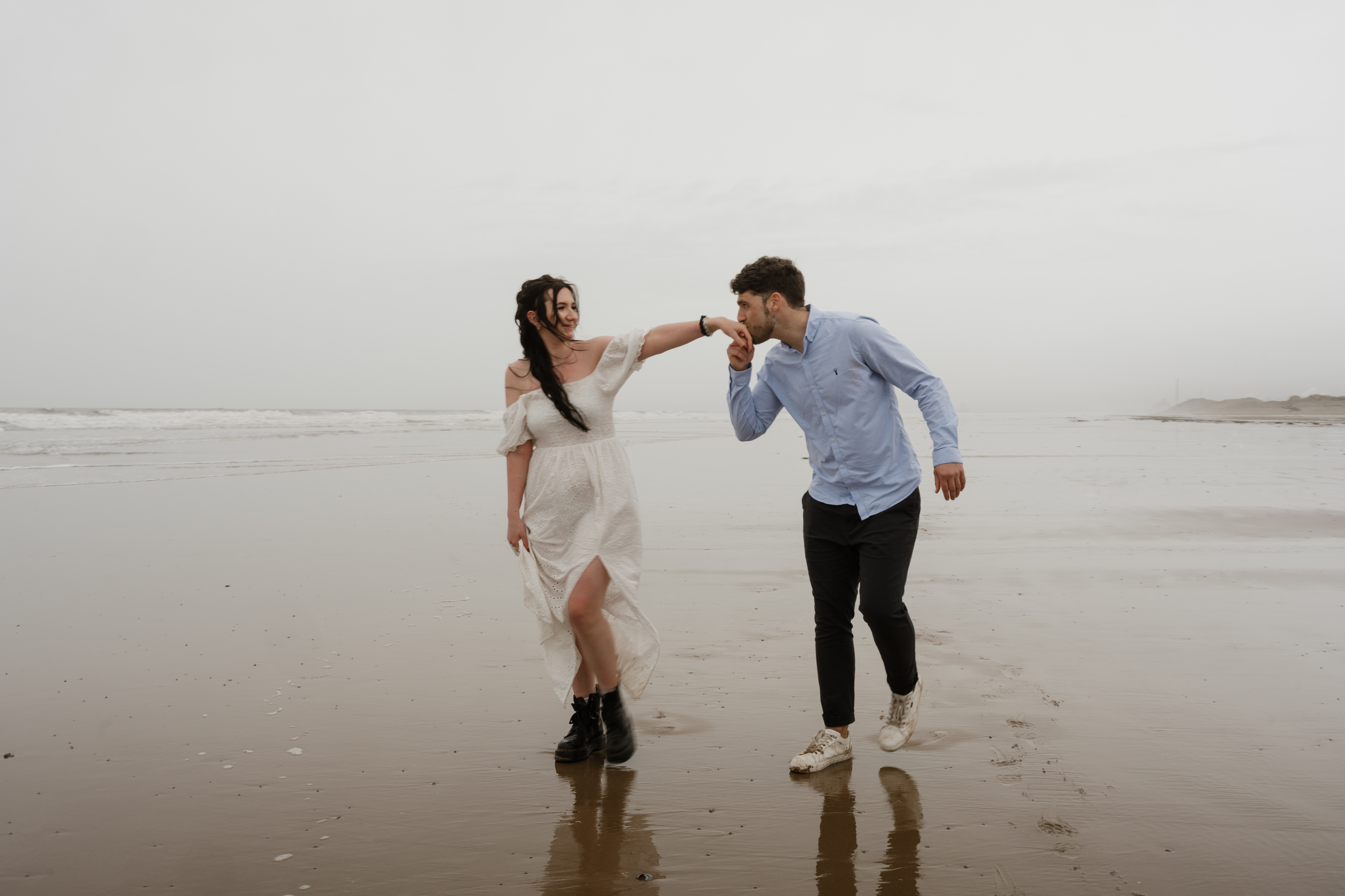 A woman wearing a white dress and a man in a blue shirt and black pants are walking on the beach. The man takes her hand and kisses it. They look happy.