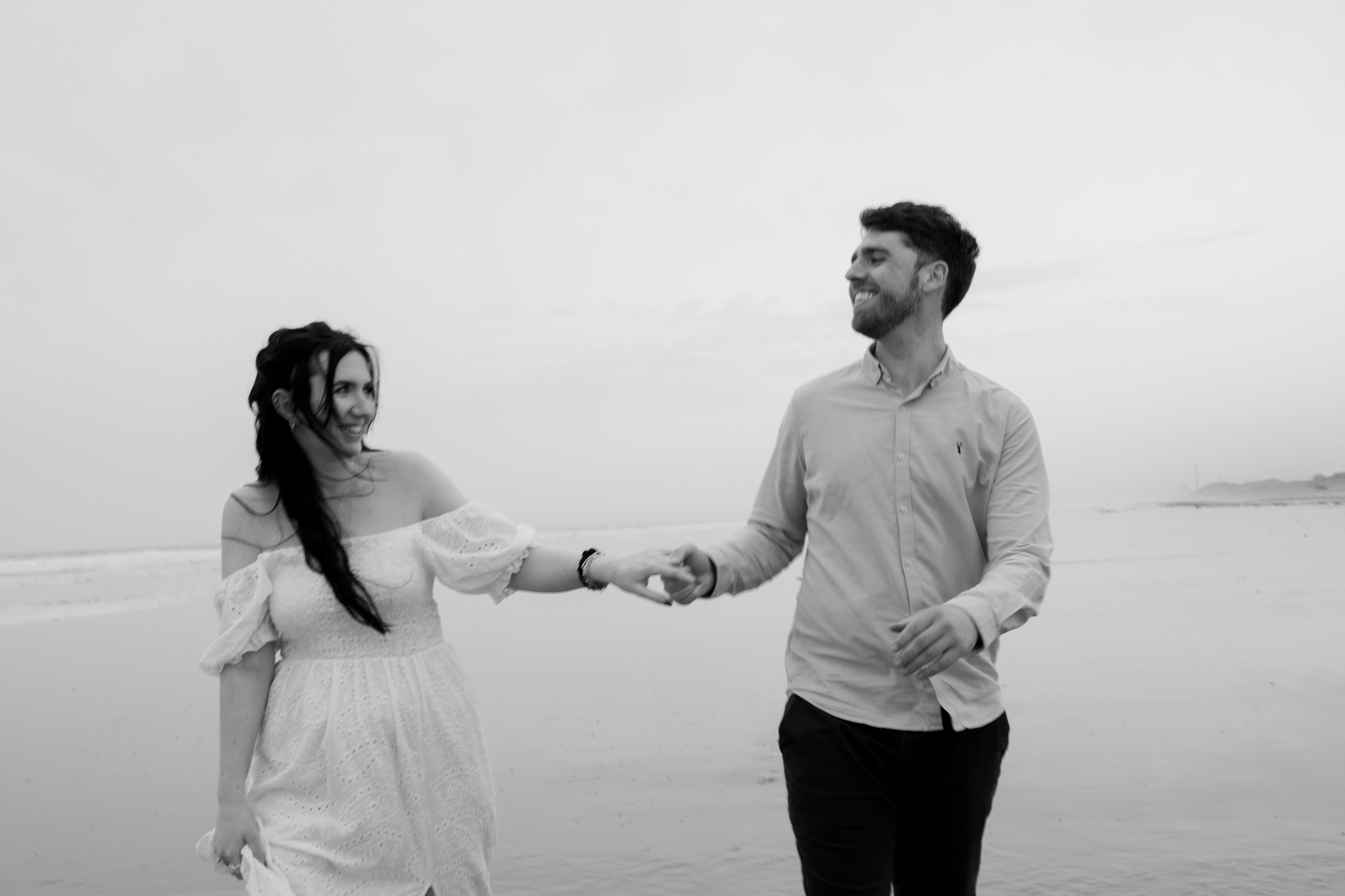 A couple is walking hand in hand along a beach, both smiling, with a calm ocean behind them. The picture is in black and white.