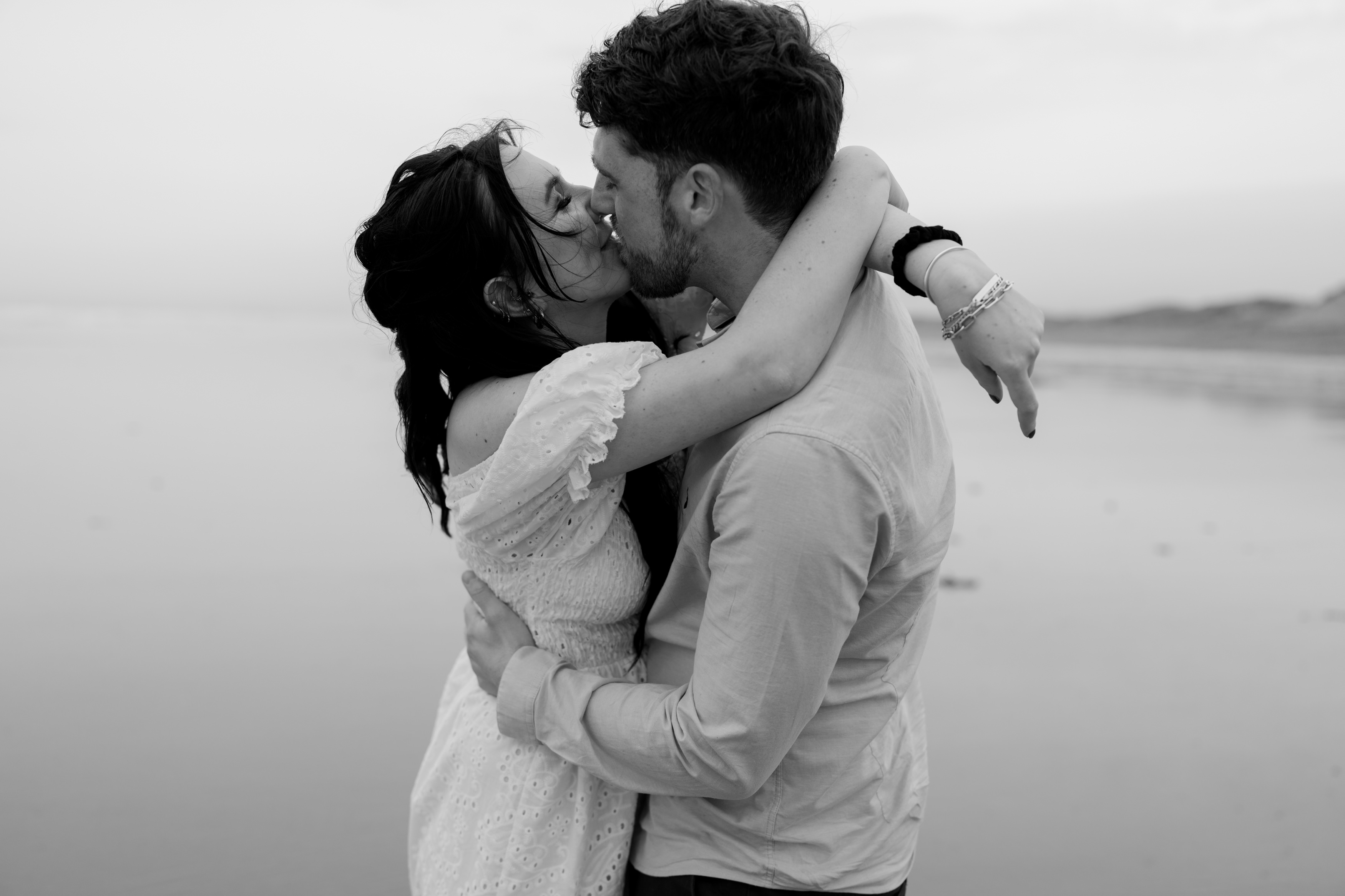 A couple is hugging and kissing on the beach, with the ocean and sky behind them. They're wearing casual clothes.