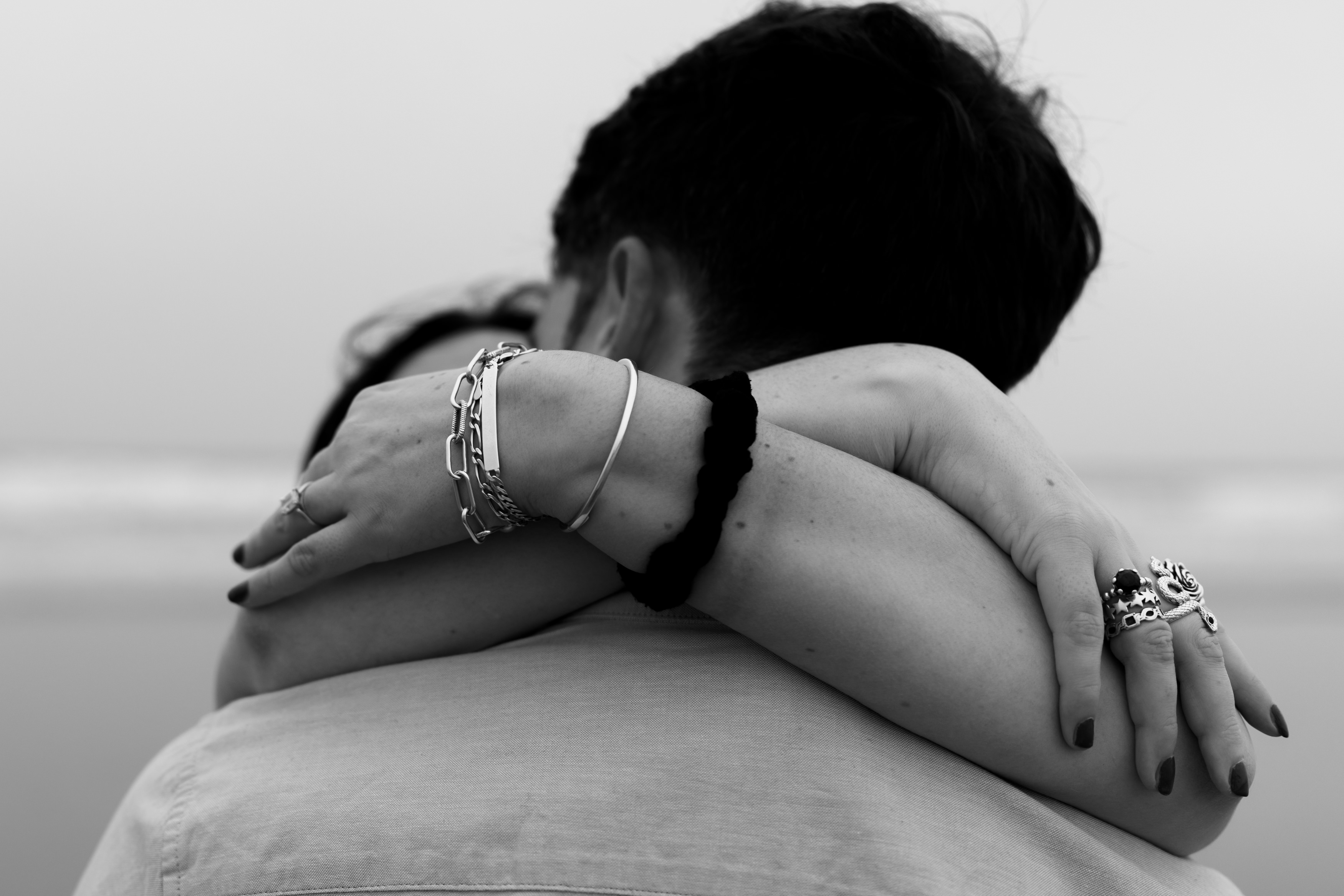 Someone wearing bracelets and rings is hugging another person from behind, while both look out at a blurry view of the sea.