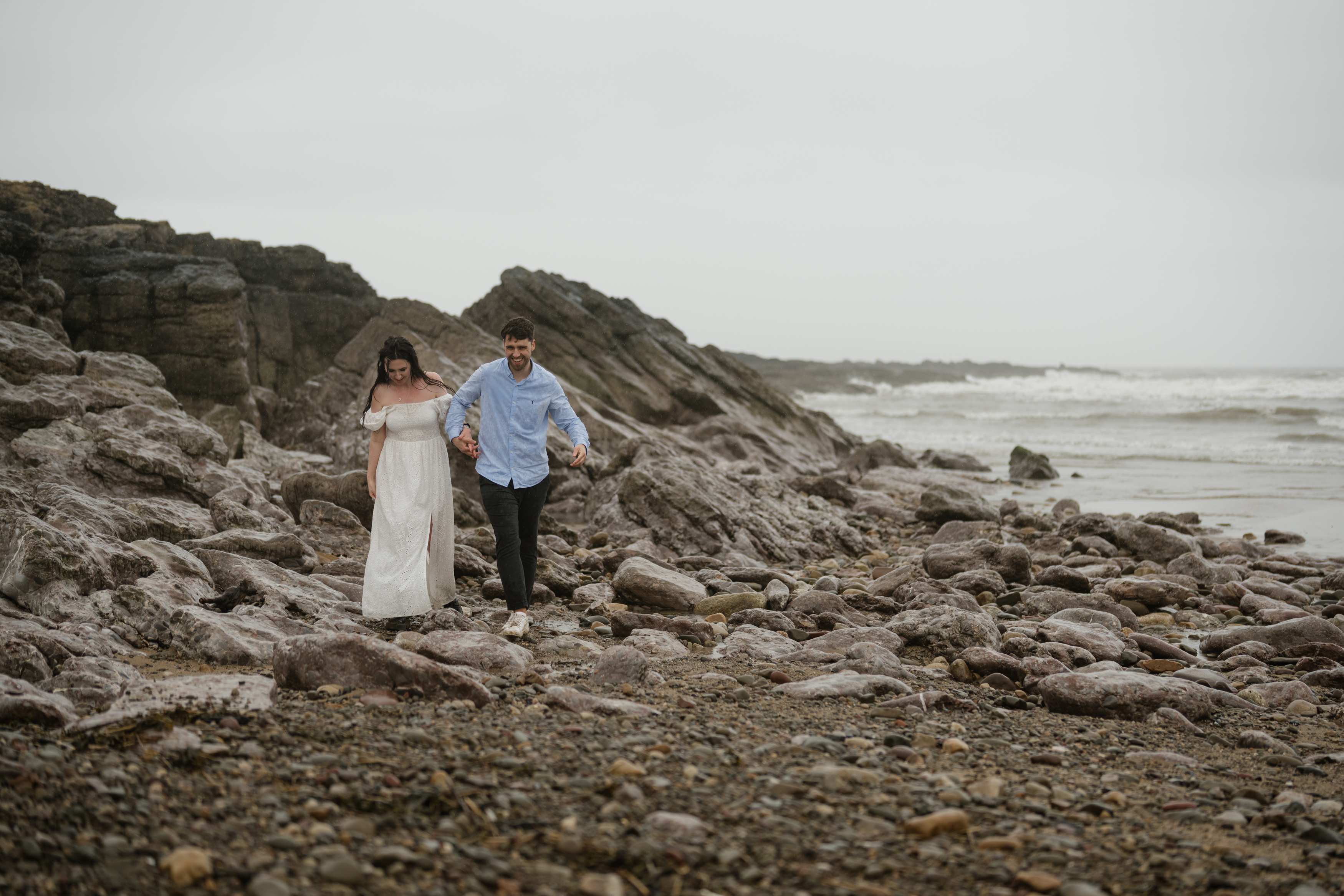A couple is holding hands as they walk on a rocky beach, with waves crashing and a cloudy sky behind them.