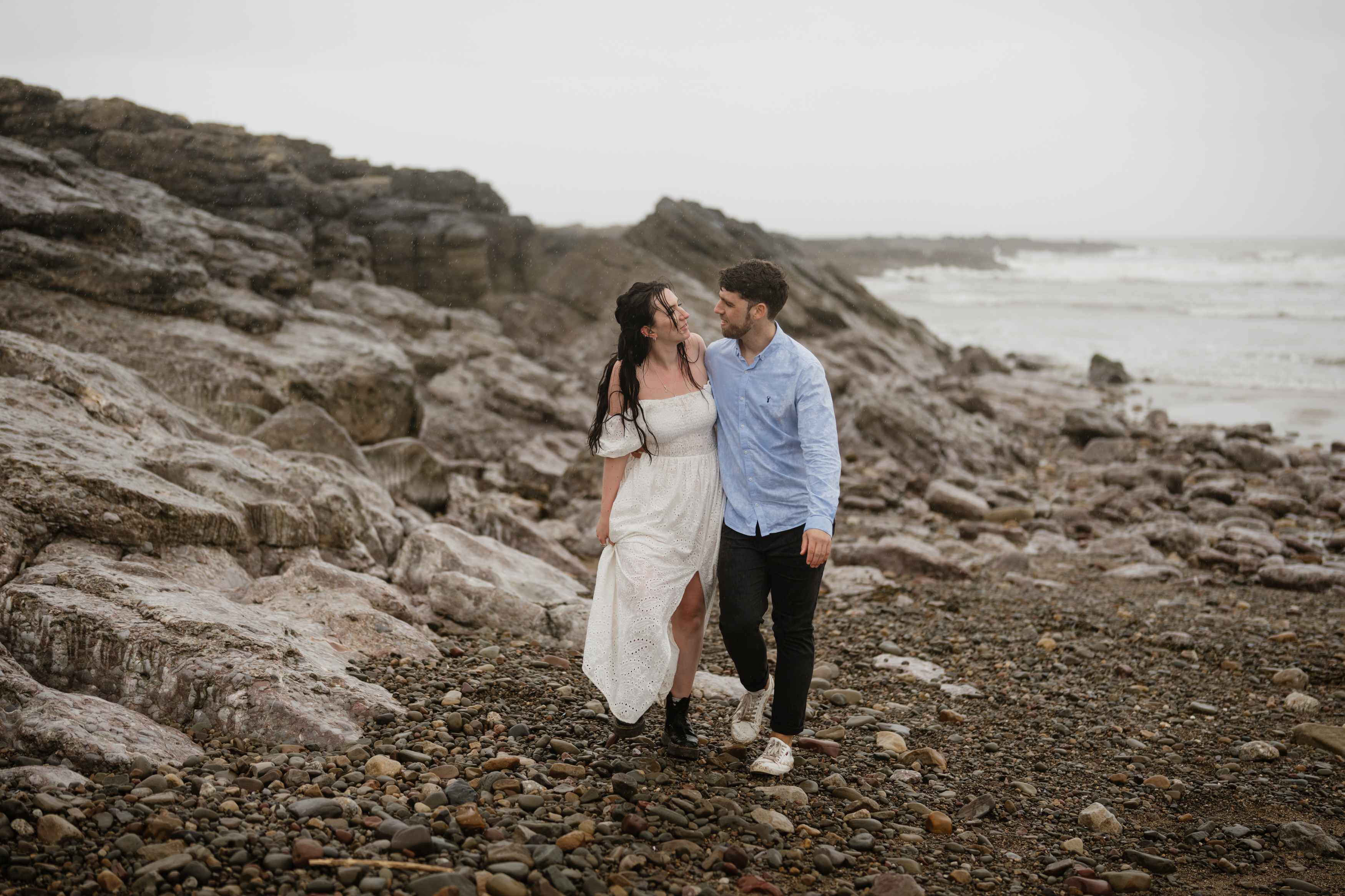 A couple is strolling along a rocky beach, dressed in casual clothes. You can see the ocean in the background beneath a cloudy sky.