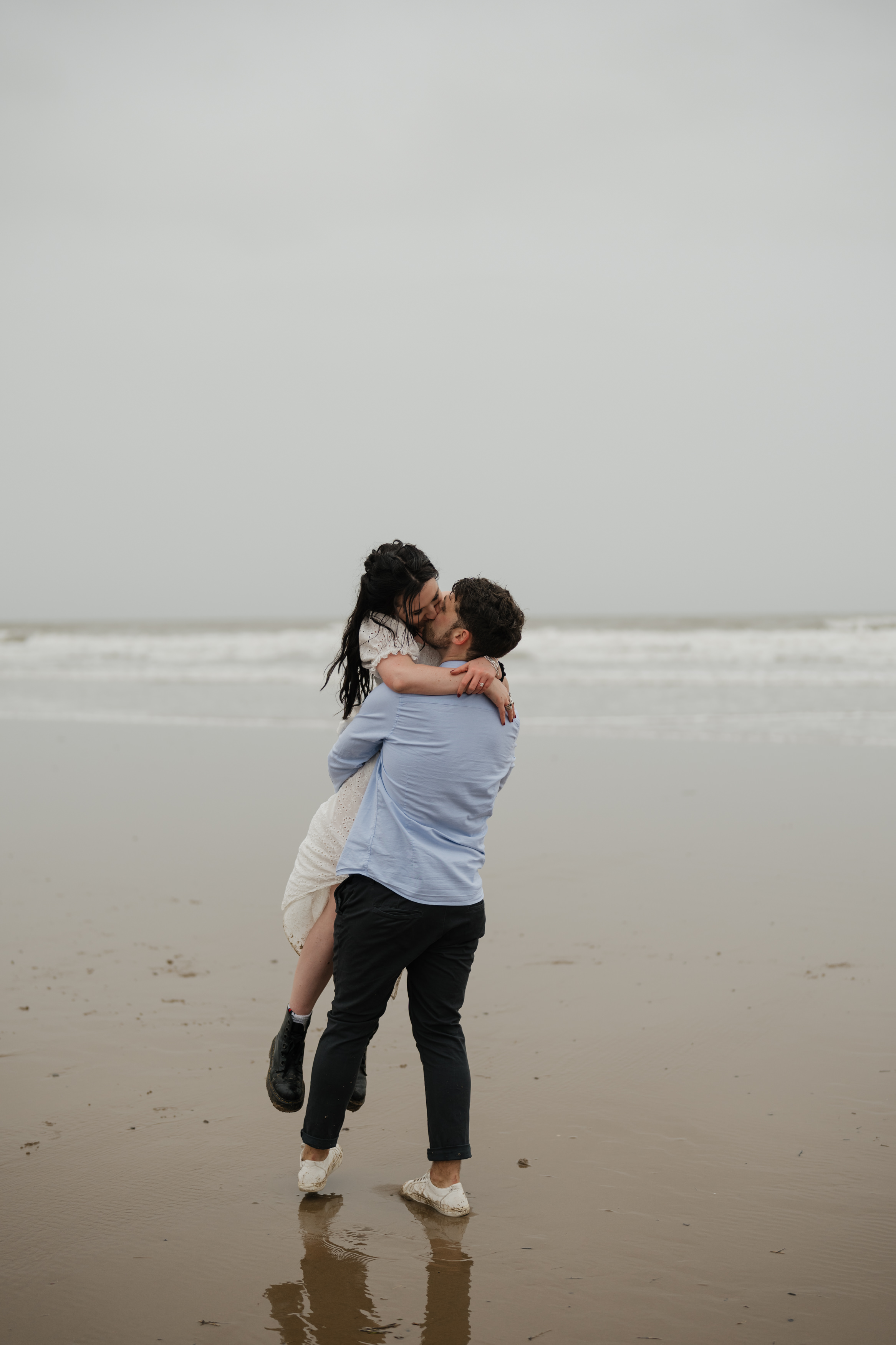 Someone is lifting another person up on a sandy beach beside the ocean, with clouds overhead.