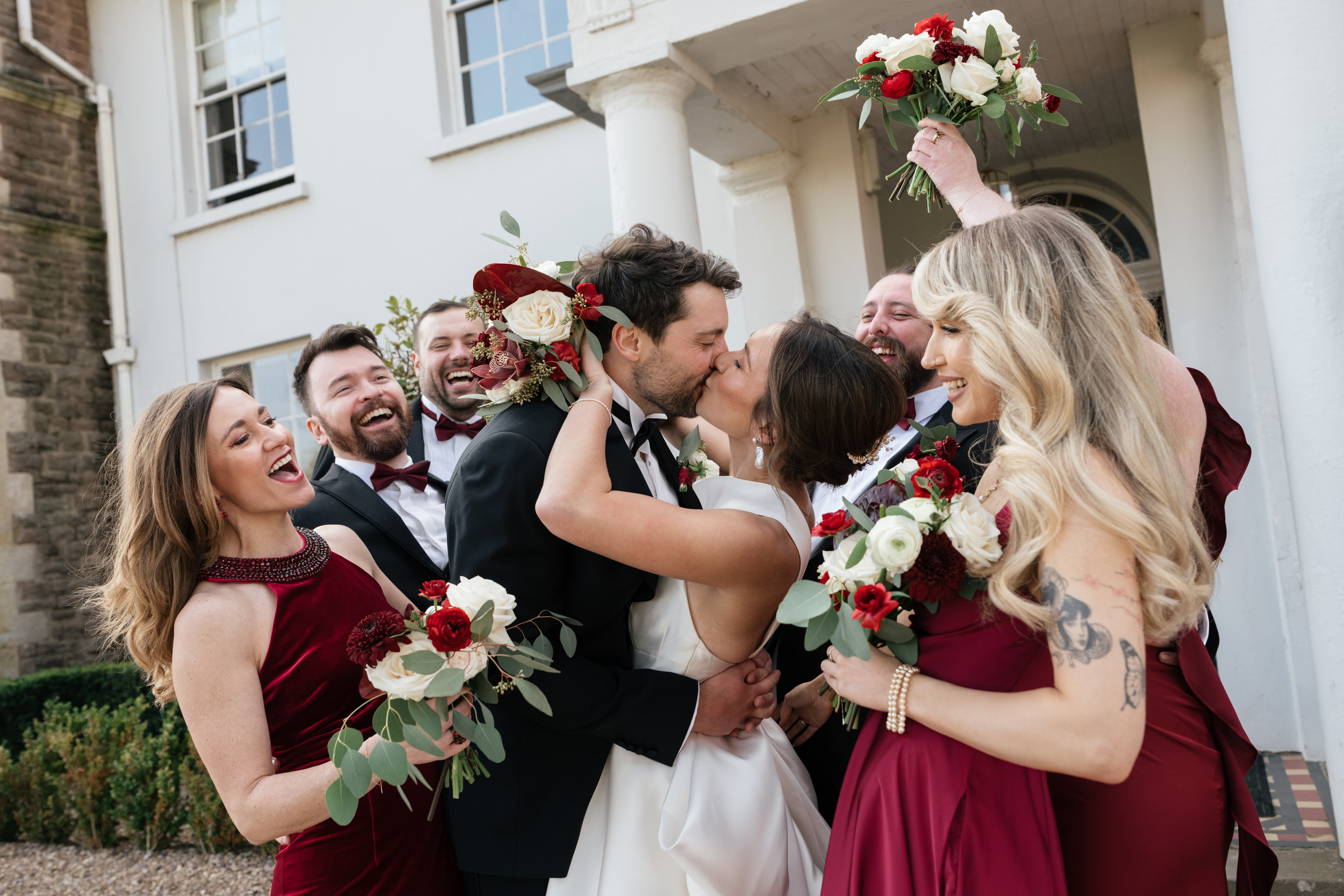 A bride and groom kiss in front of a building, with their happy wedding party around them dressed in burgundy outfits and holding red and white flower bouquets.