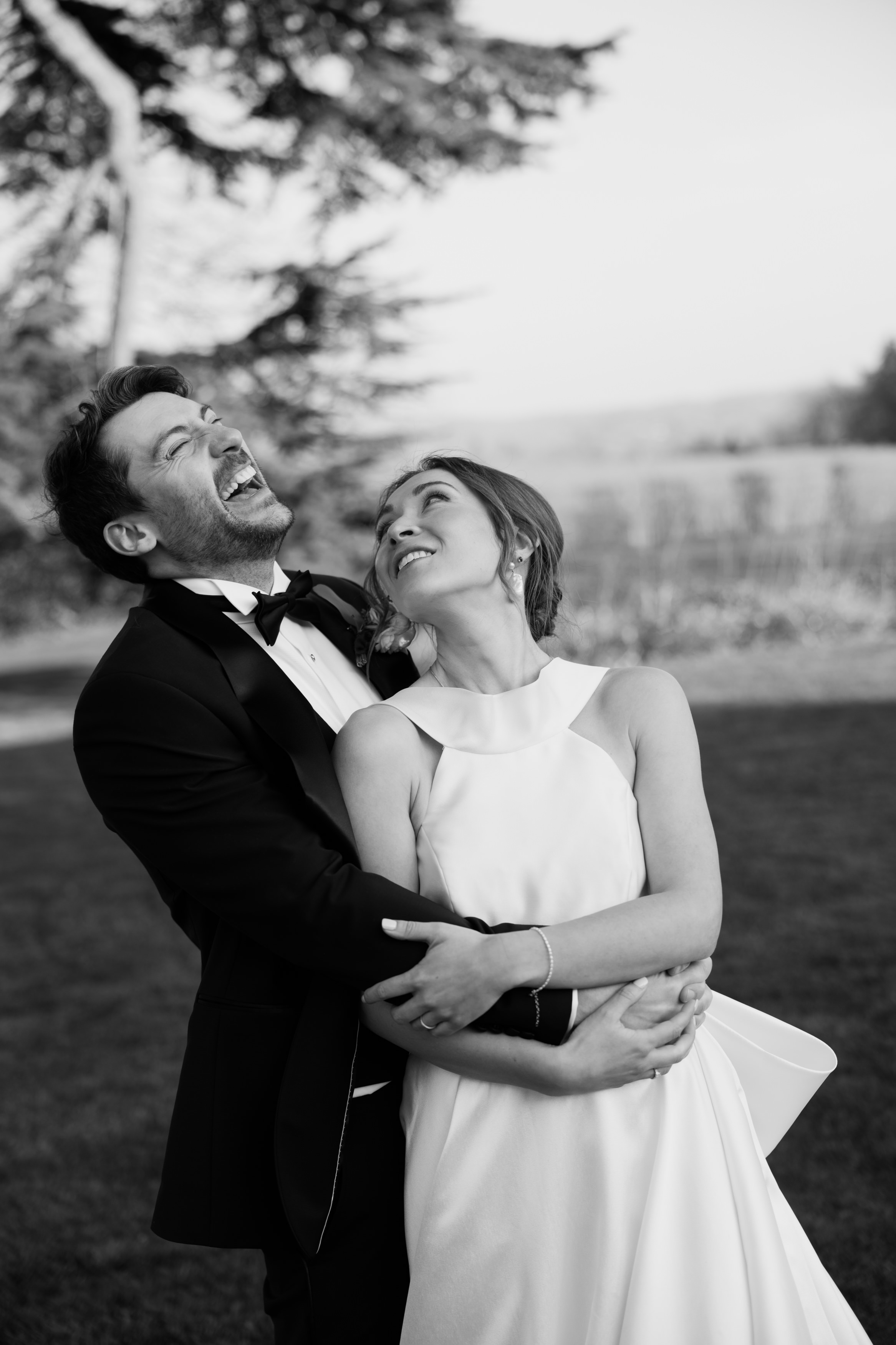 A couple dressed up in formal clothes stands outside, hugging and laughing together, with trees and open countryside behind them.