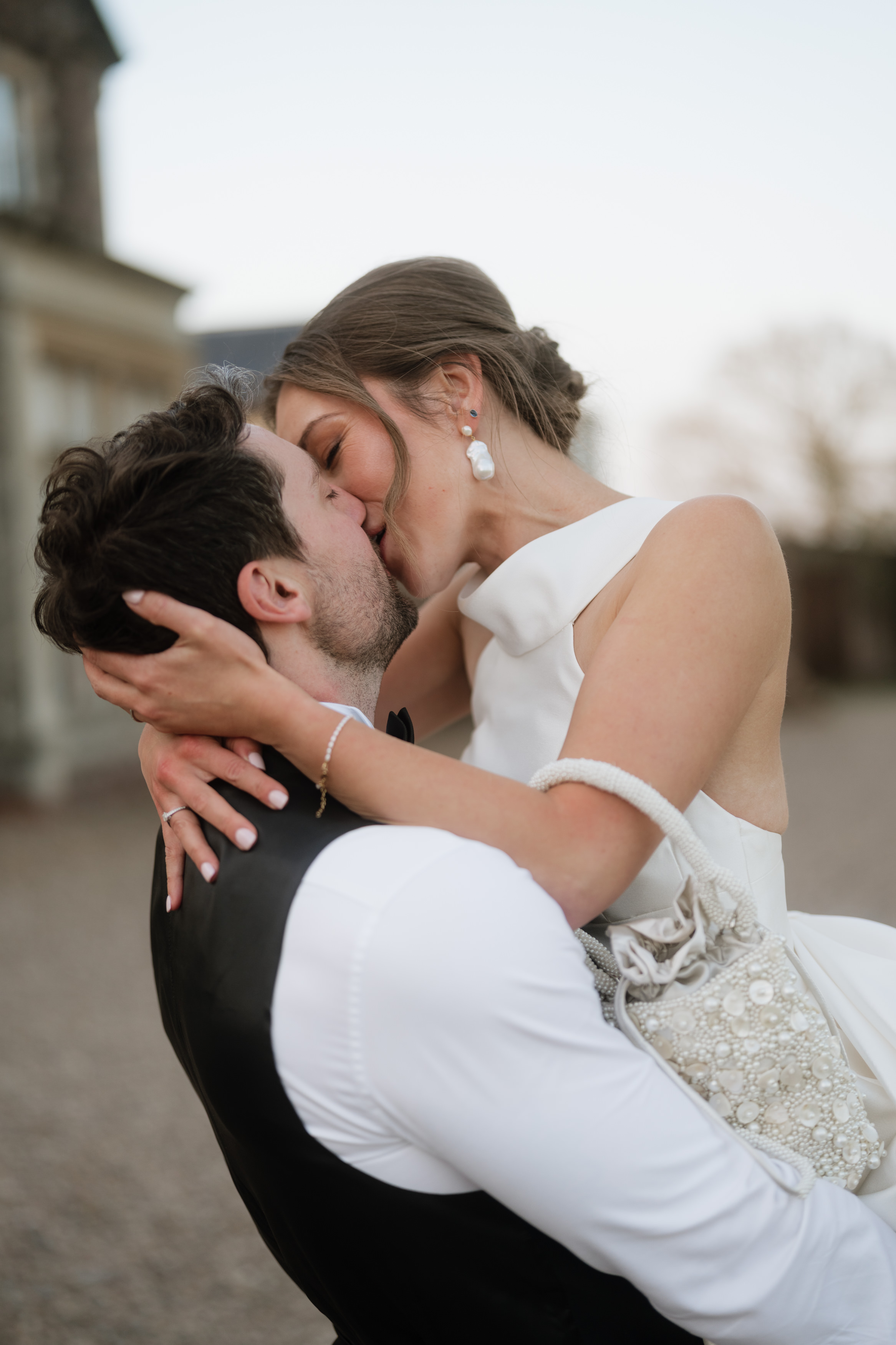 A couple in wedding clothes kisses outside—one is wearing a white dress, and the other has on a white shirt with a black waistcoat.