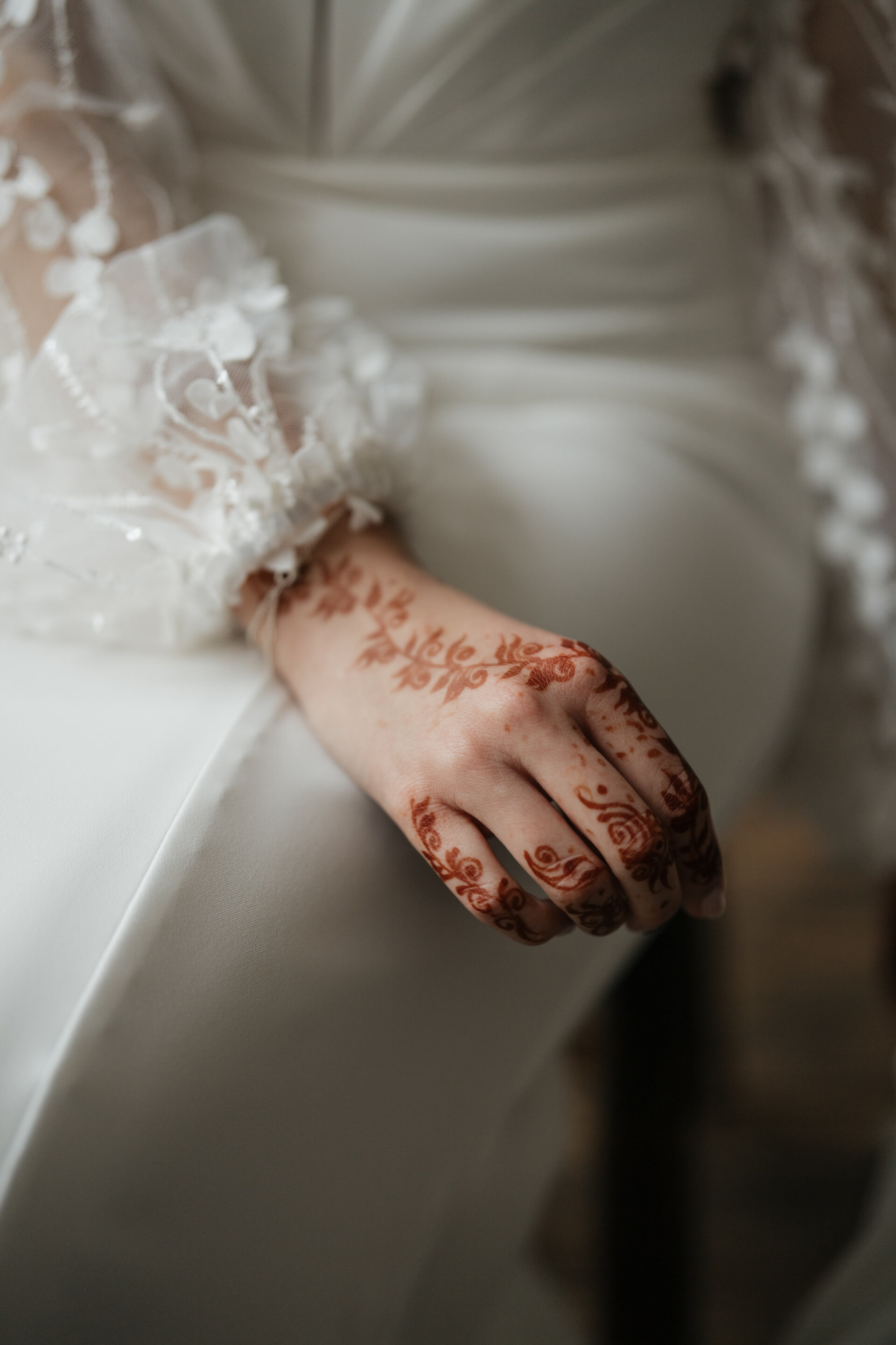 A hand with detailed henna designs and lace sleeves rests on a white dress.