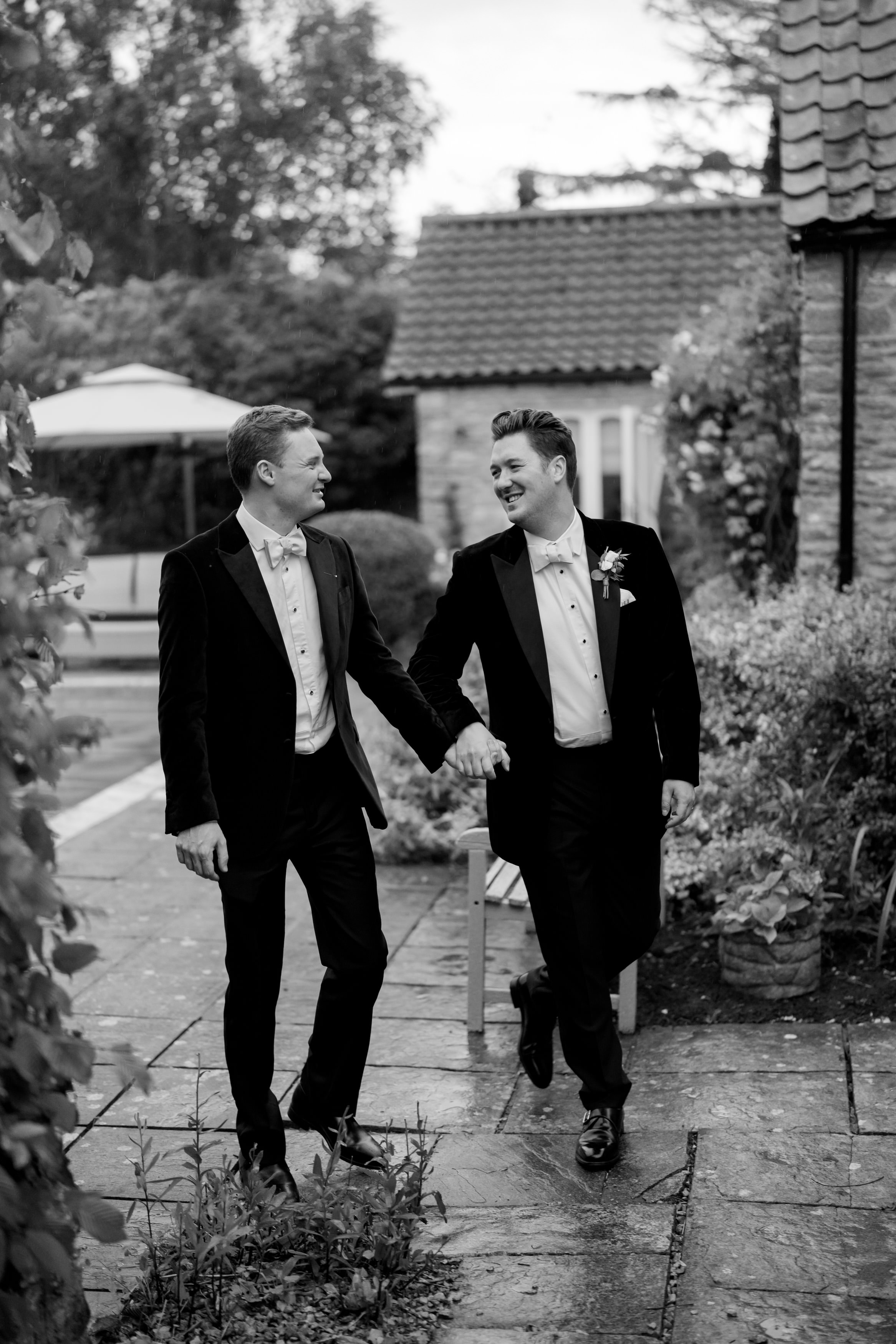 Two people dressed up, holding hands and smiling as they walk on a stone path outside.