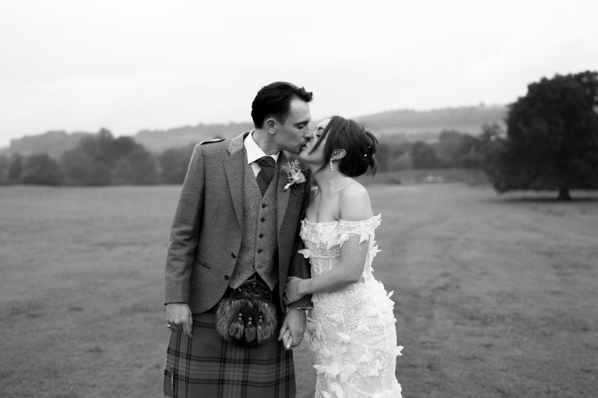A couple dressed up in fancy clothes is kissing outside. The man has on a kilt, and the woman is wearing a white dress. They're surrounded by a grassy field.
