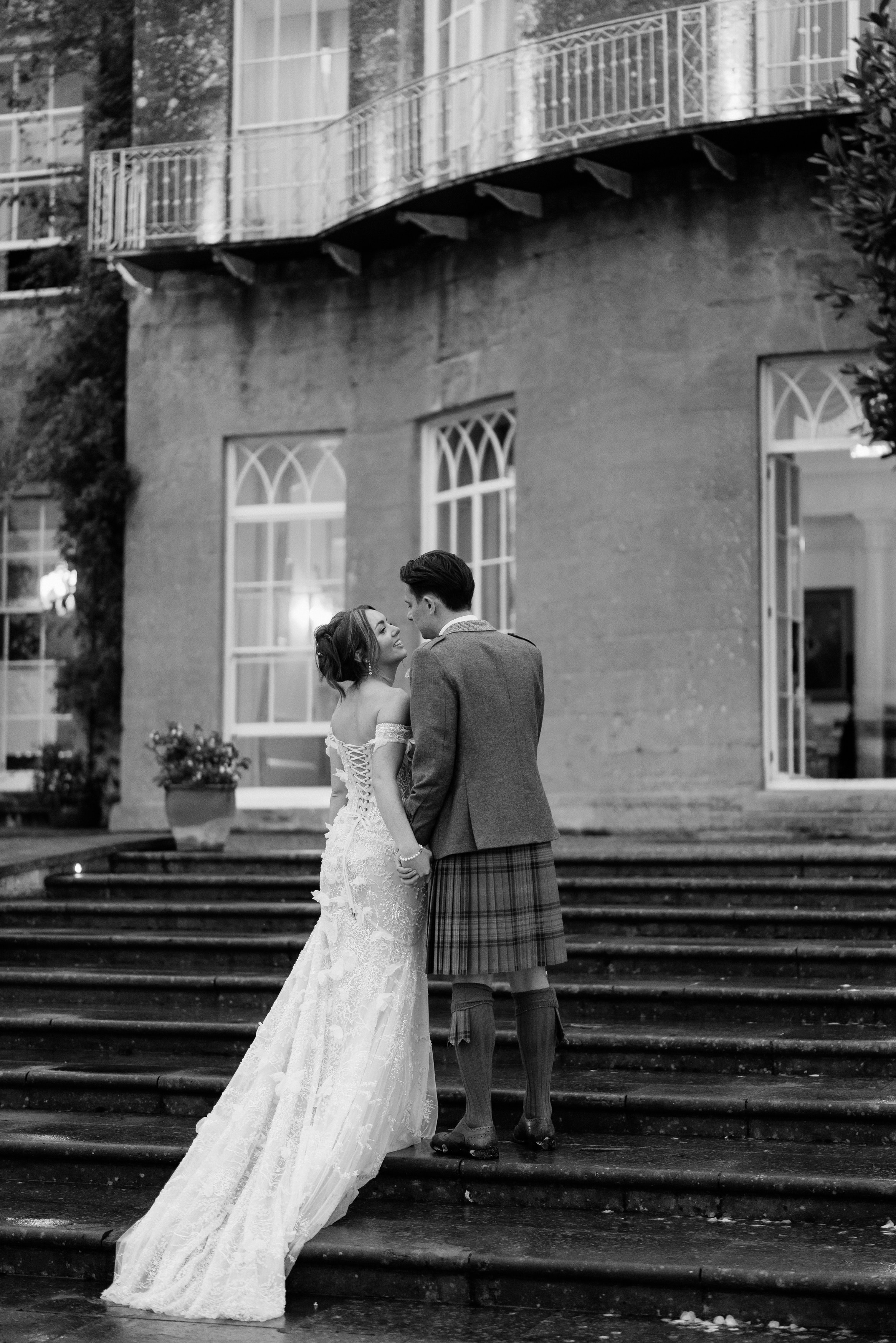 A bride in a long dress and a groom in a kilt take a moment together on the steps outside of a big building.