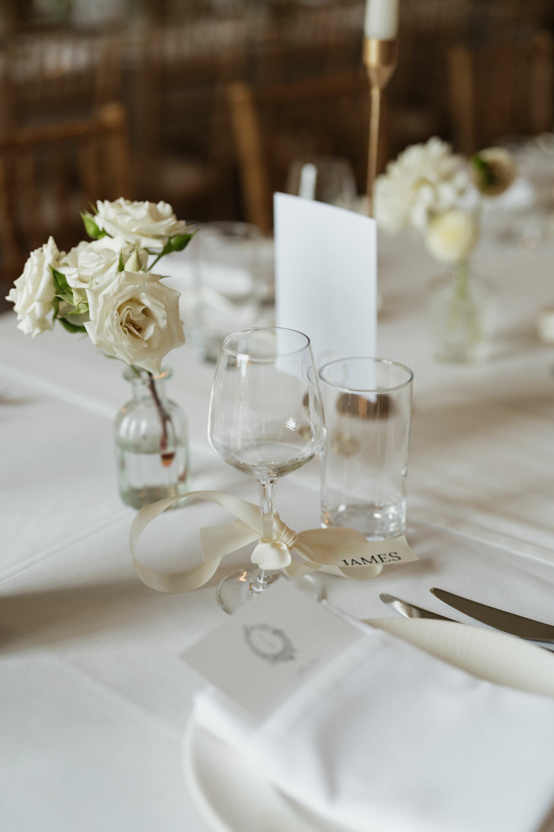 A nicely set table with white roses in a vase, an empty wine glass, and a name card on a white tablecloth.