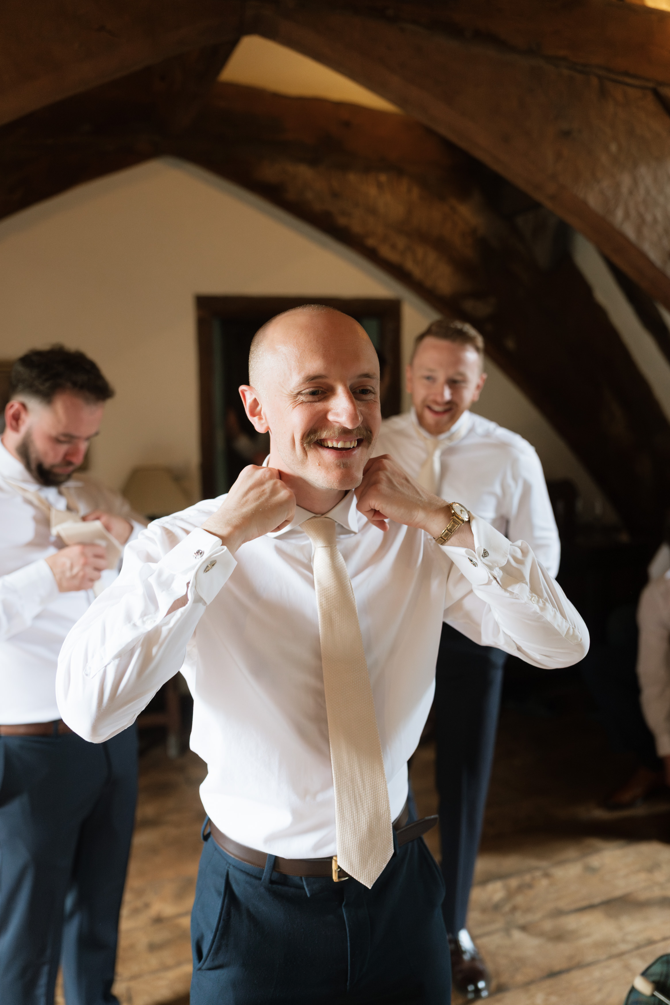 A man wearing a white shirt and tie smiles as he fixes his collar, while two other men dressed the same are getting ready in a rustic-looking room.