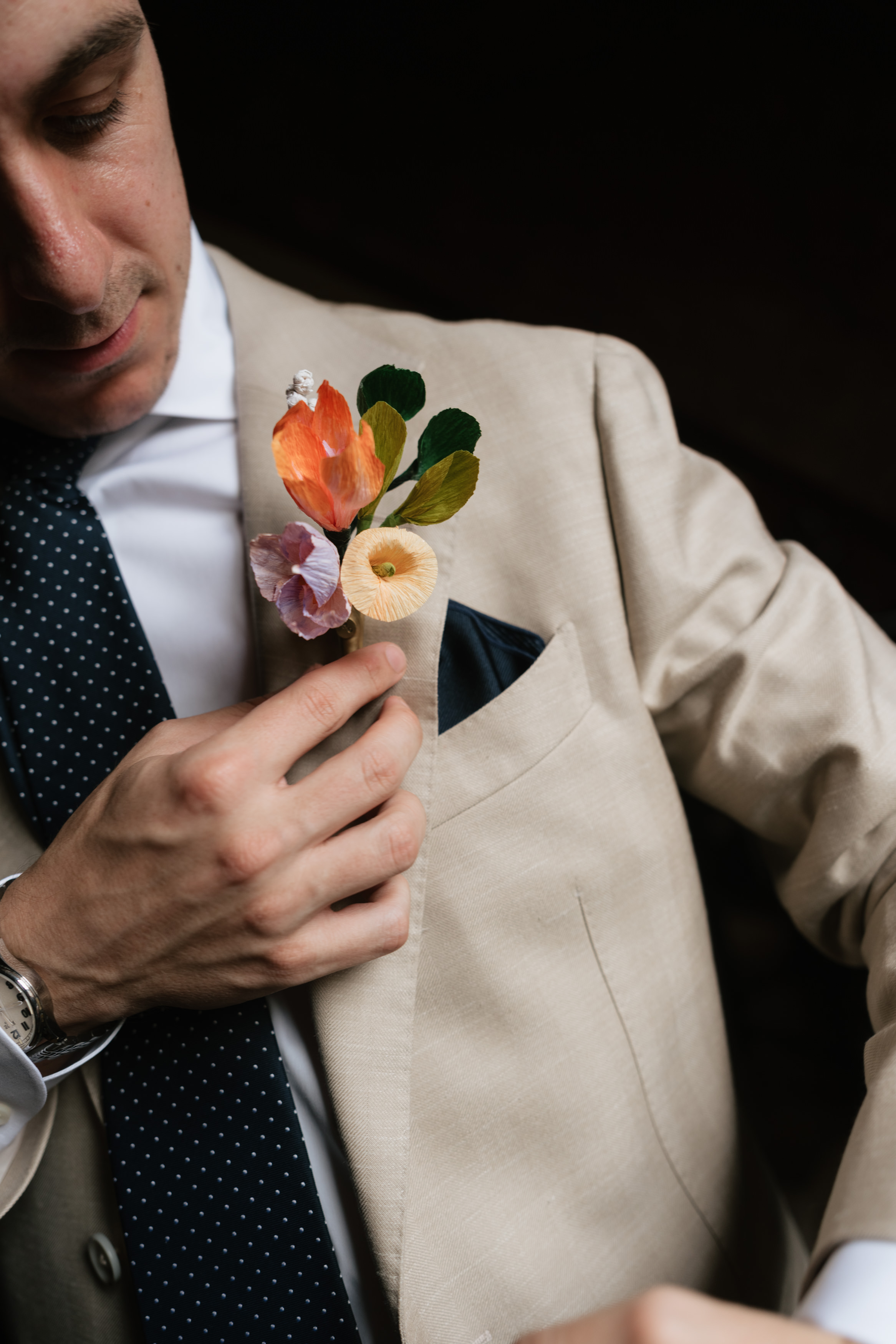 A man wearing a beige suit jacket and a spotted tie is fixing a colourful buttonhole on his lapel, with a dark background behind him.