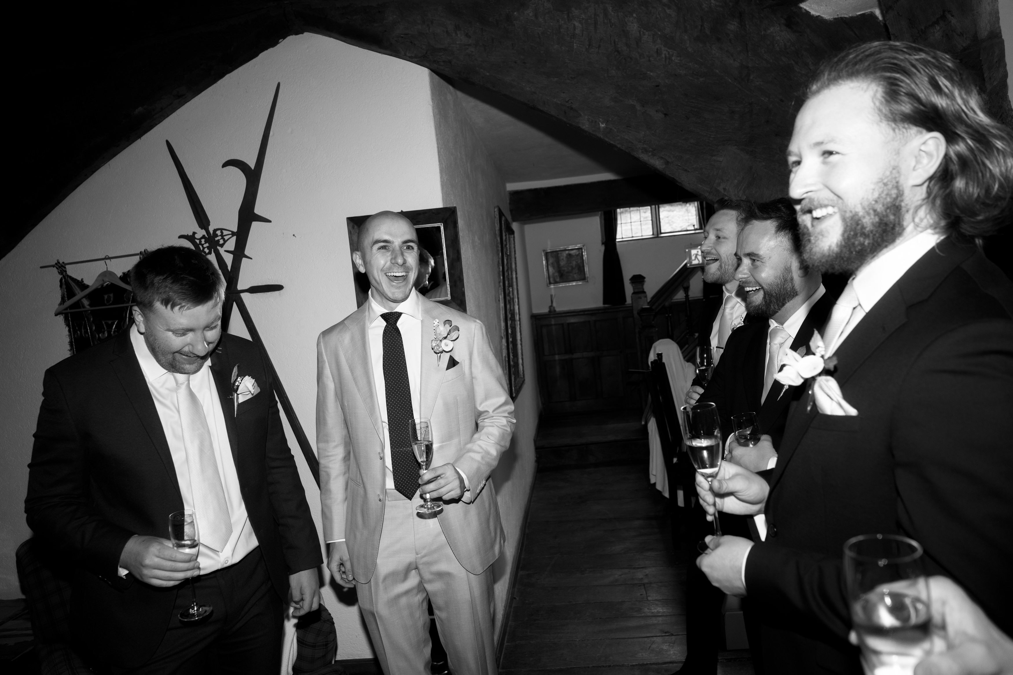Five men dressed in suits are inside a room with wooden accents, some of them holding champagne glasses. They’re smiling and laughing together, clearly enjoying themselves.