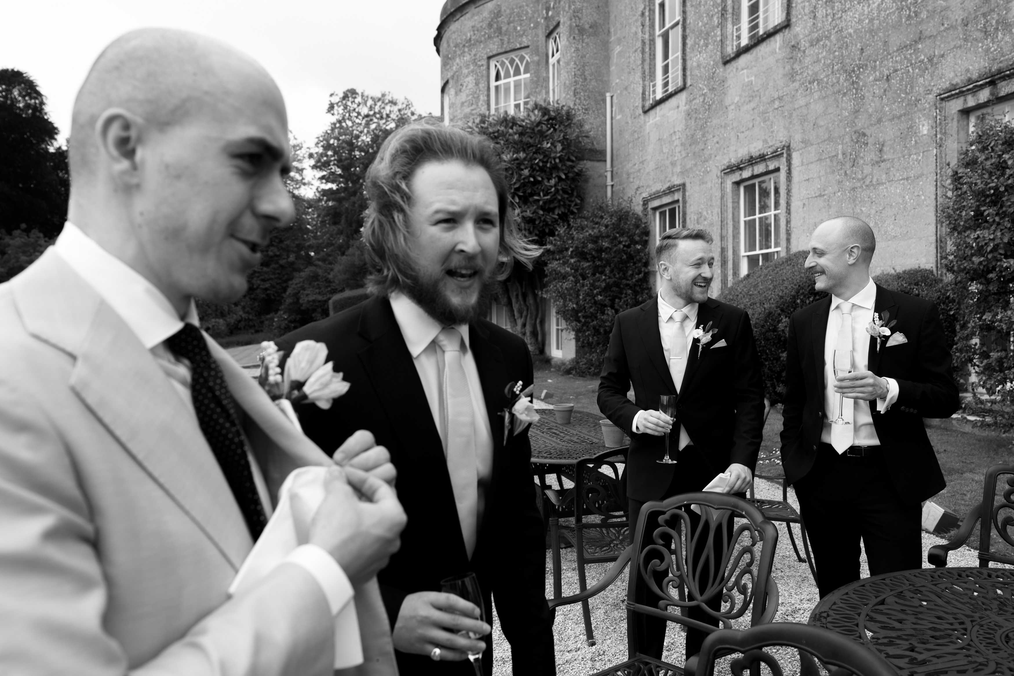 Four men wearing suits are talking and laughing together outside a large stone building. They’re standing near some metal tables and chairs.