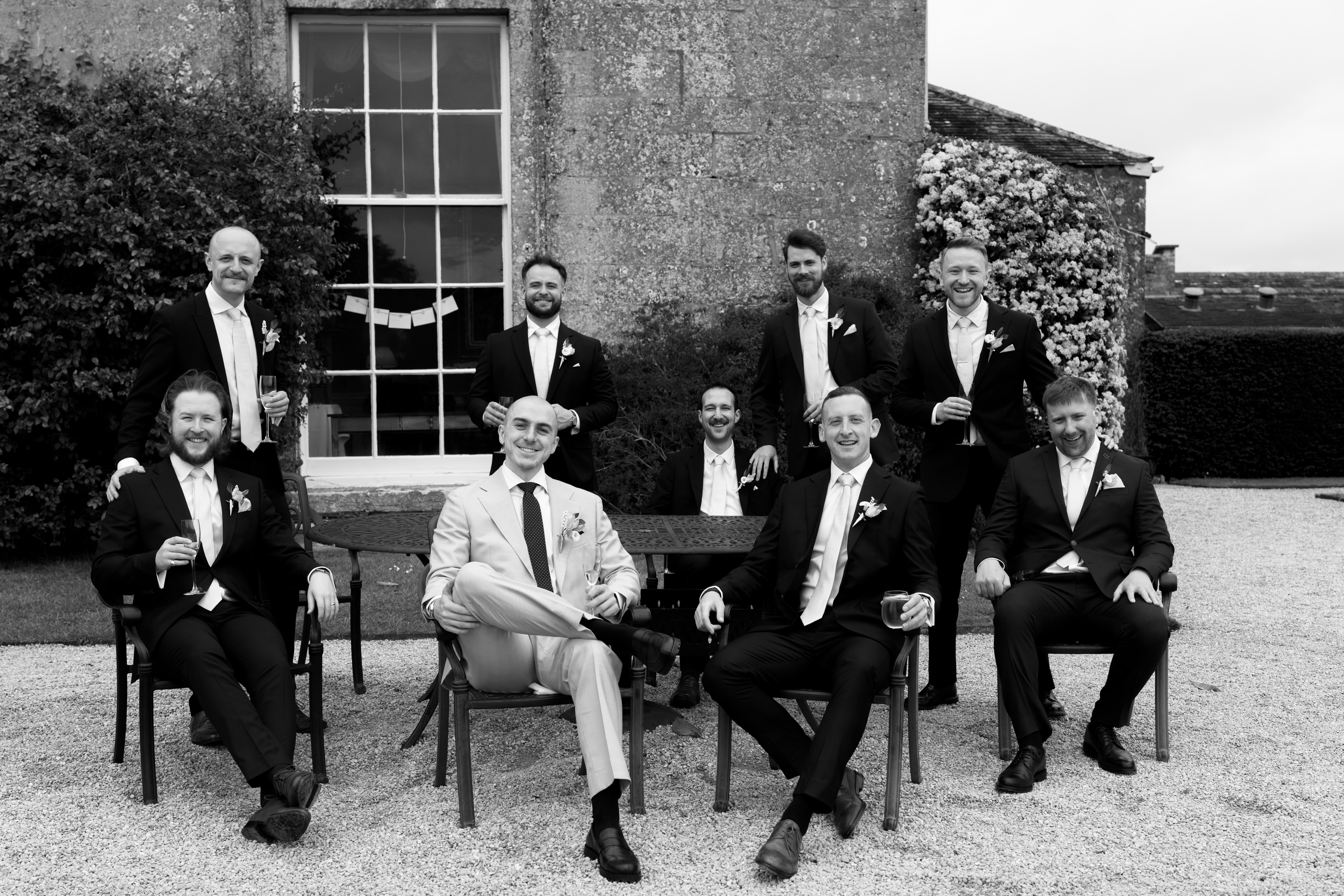 Eight men wearing suits, with some sitting and others standing, are outside a stone building. They all have drinks in their hands and are smiling at the camera.