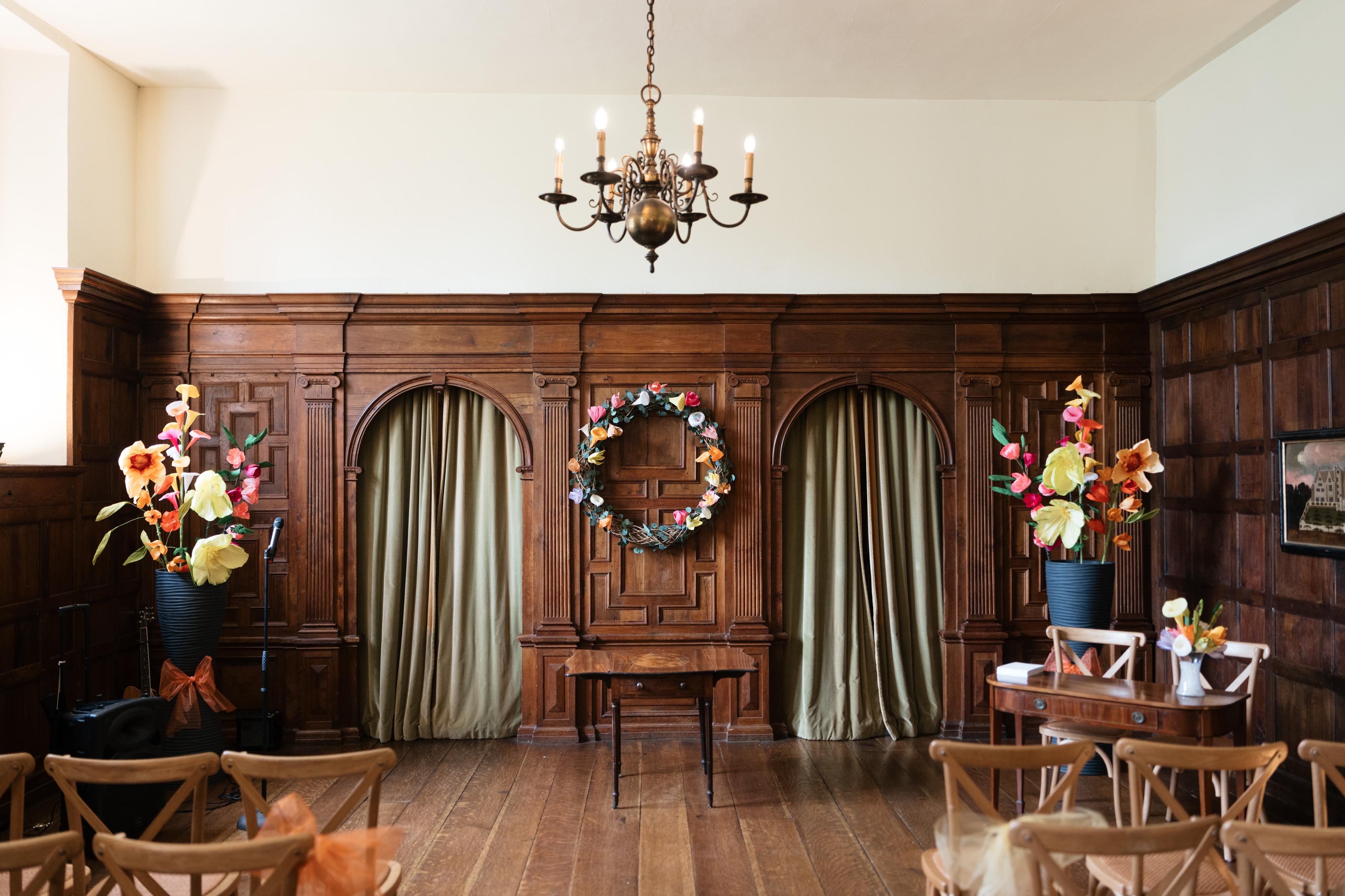 A room with wood-panelled walls, a chandelier hanging from the ceiling, a wreath in the centre of the wall, and two tall vases filled with colourful flowers. There are rows of chairs set up facing a small table and bench at the front.