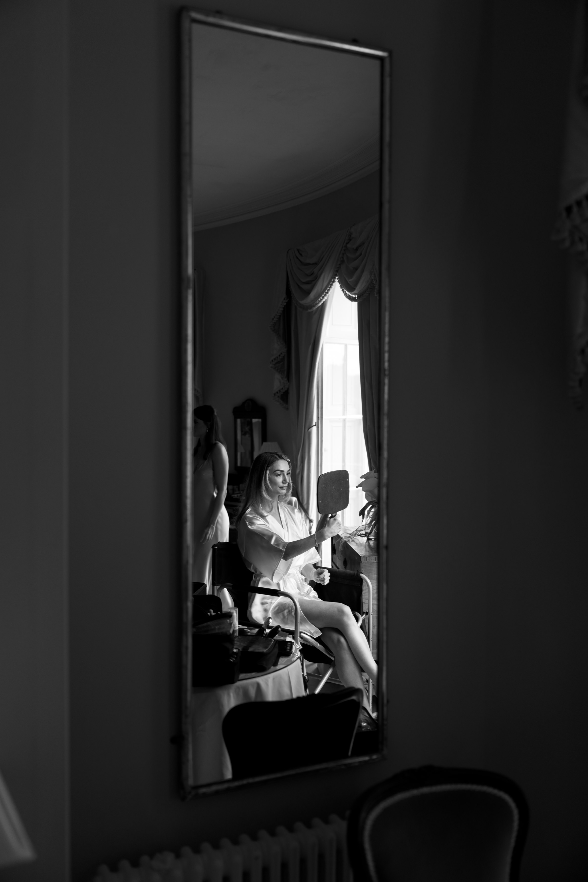 A woman is sitting in front of a mirror, holding a small hand-held mirror. You can see her reflection in a large wall mirror behind her in a softly lit room.