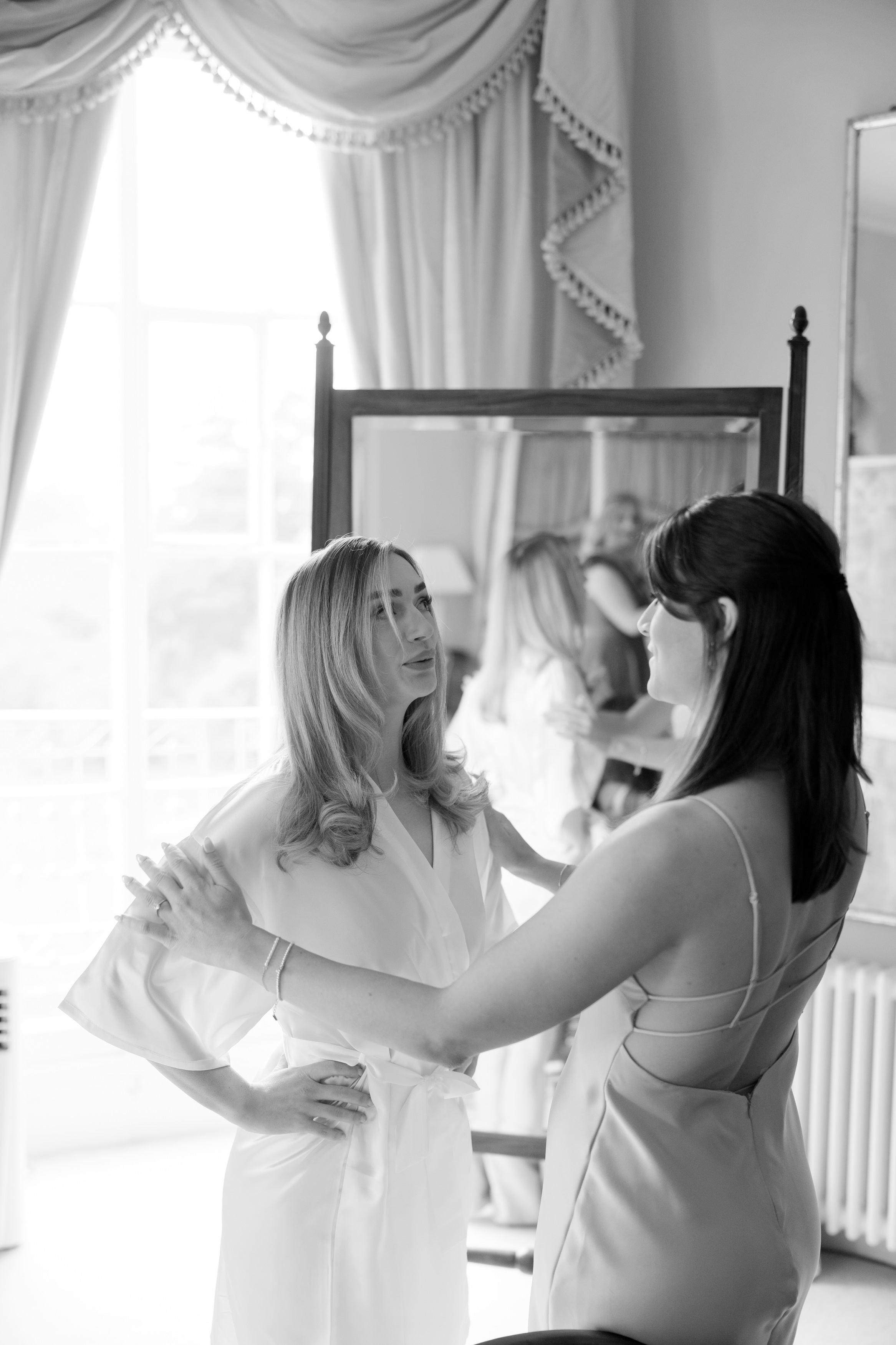 Two women are indoors, standing and facing each other. One is dressed in a dressing gown and the other in a dress. There’s a mirror and a window behind them.