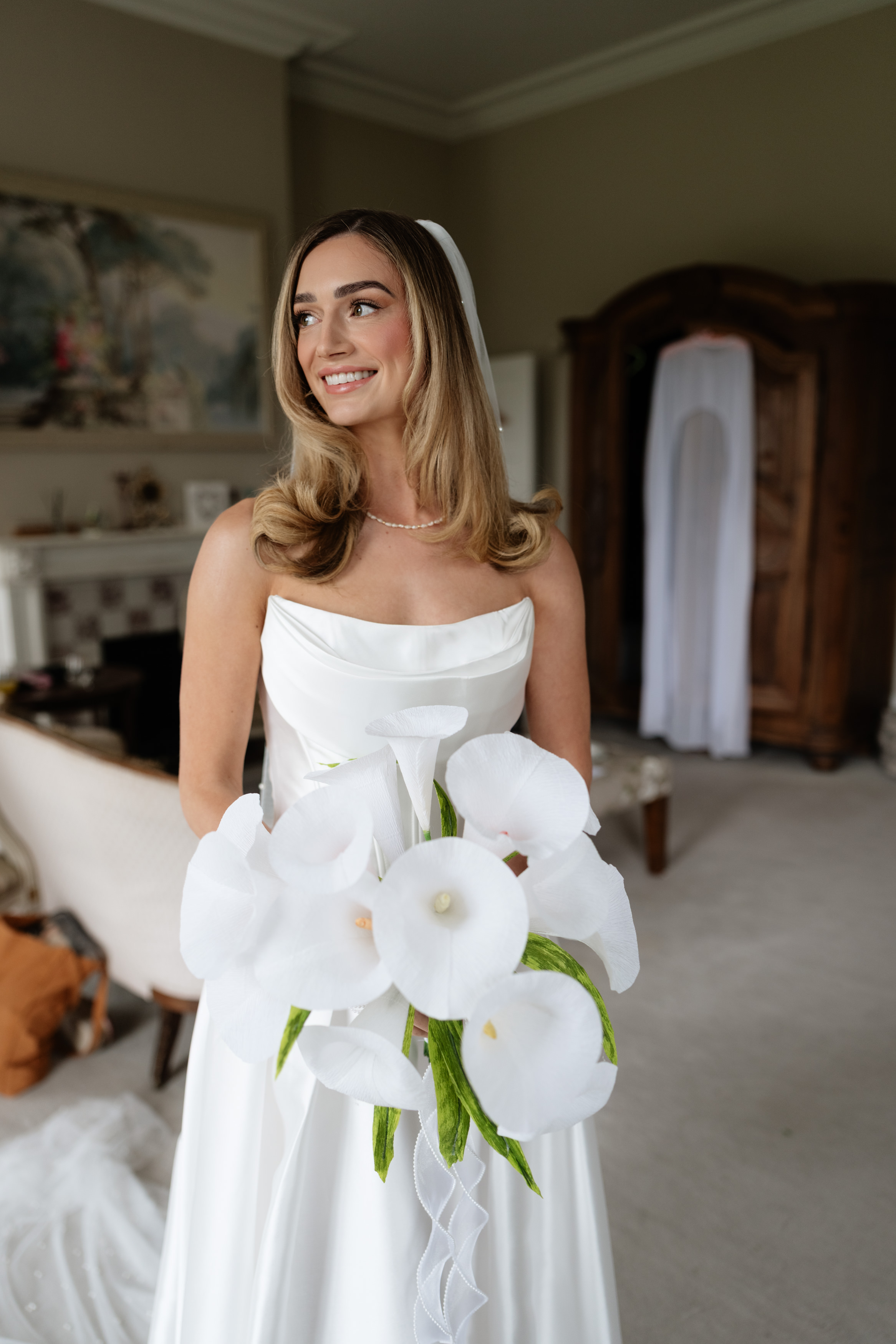 A bride wearing a white dress is holding a bunch of white flowers and smiling whilst standing in a gently lit room with traditional decorations.