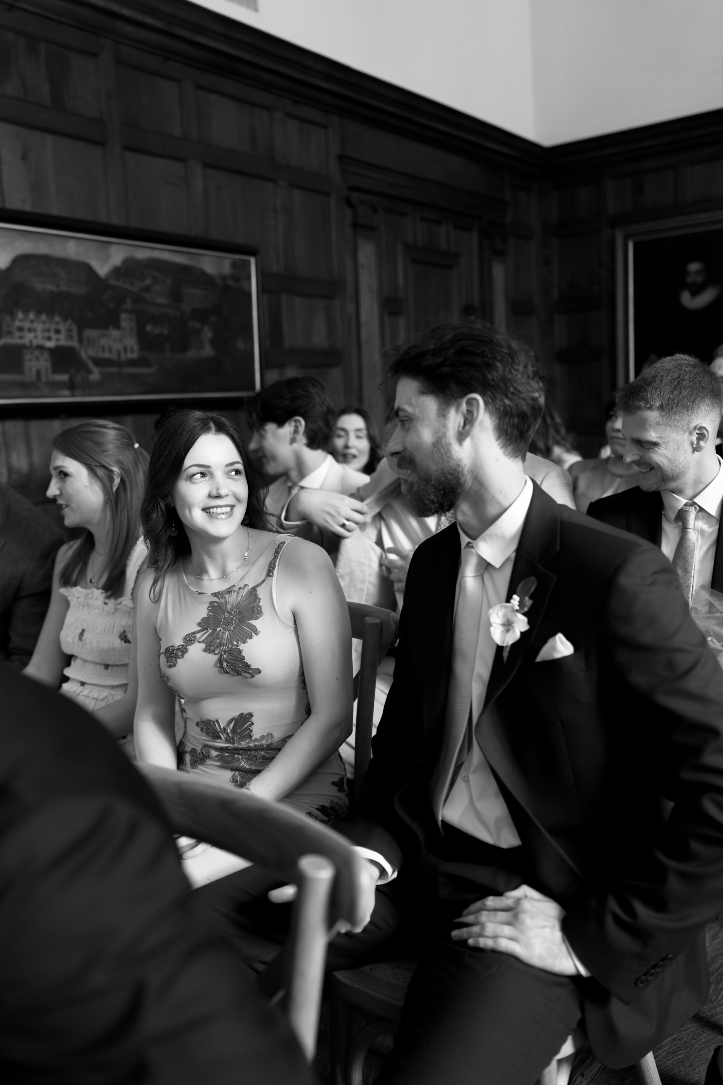 A woman wearing a floral dress and a man in a suit are sitting with other people in a room with wooden walls, smiling at each other.