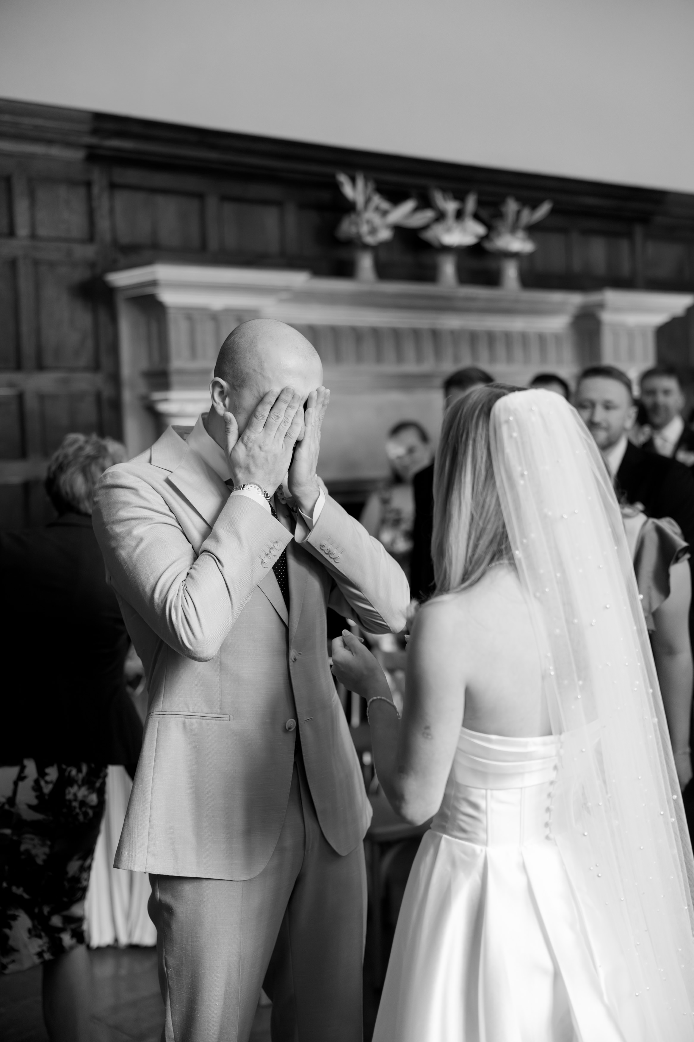 The groom hides his face in his hands as he stands in front of the bride at their wedding ceremony, surrounded by guests in a room with wood-panelled walls.