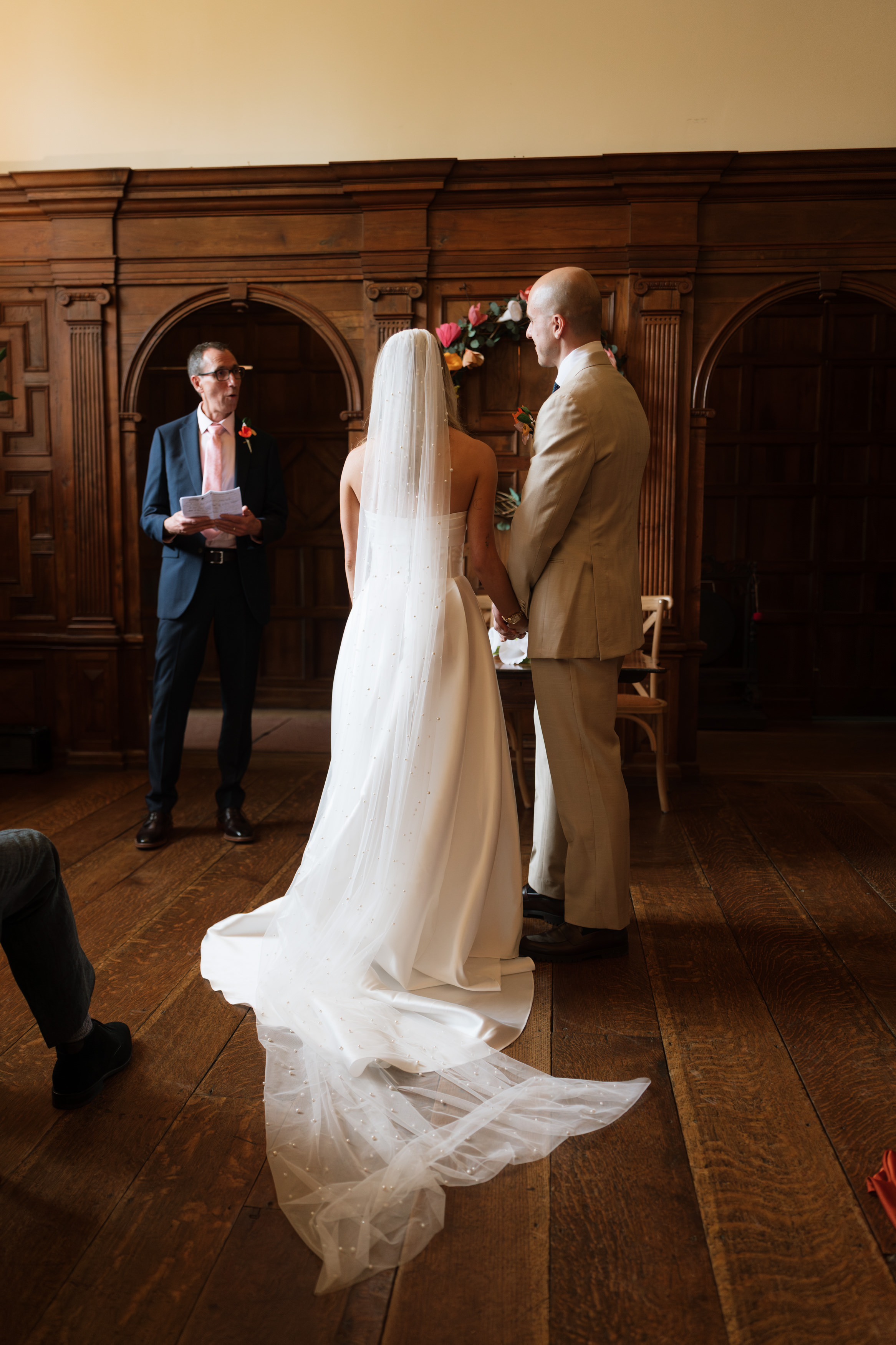 A bride and groom are standing indoors at their wedding, looking at each other, while an officiant stands next to them holding some papers.