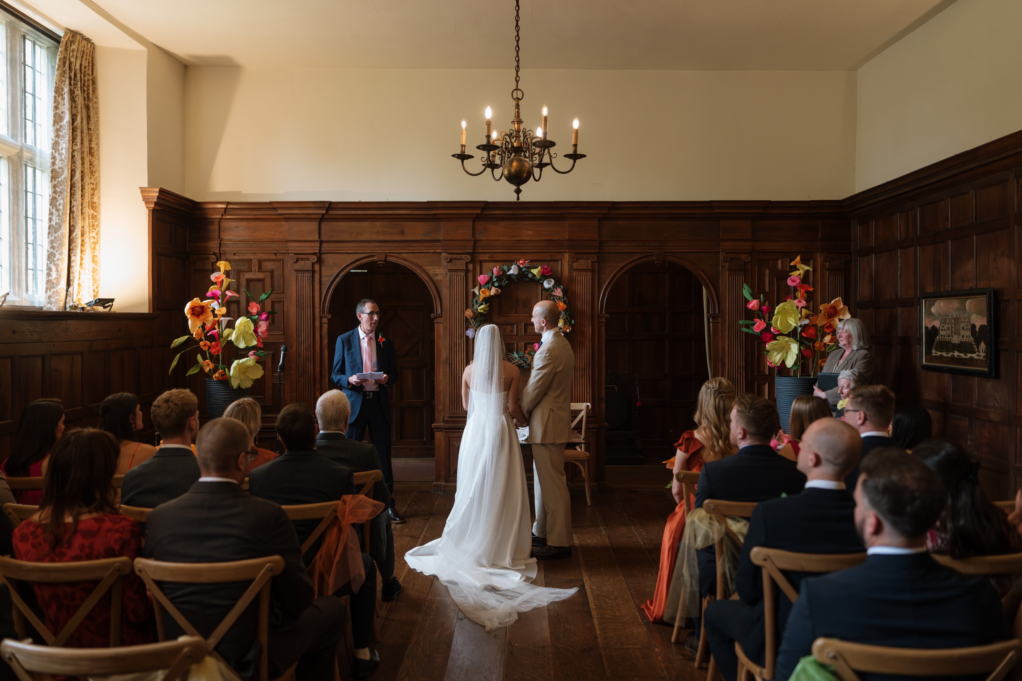 A couple stands at the front of a wood-panelled room, facing the person leading their ceremony. Guests sit around them, and there are flower decorations and a chandelier above.