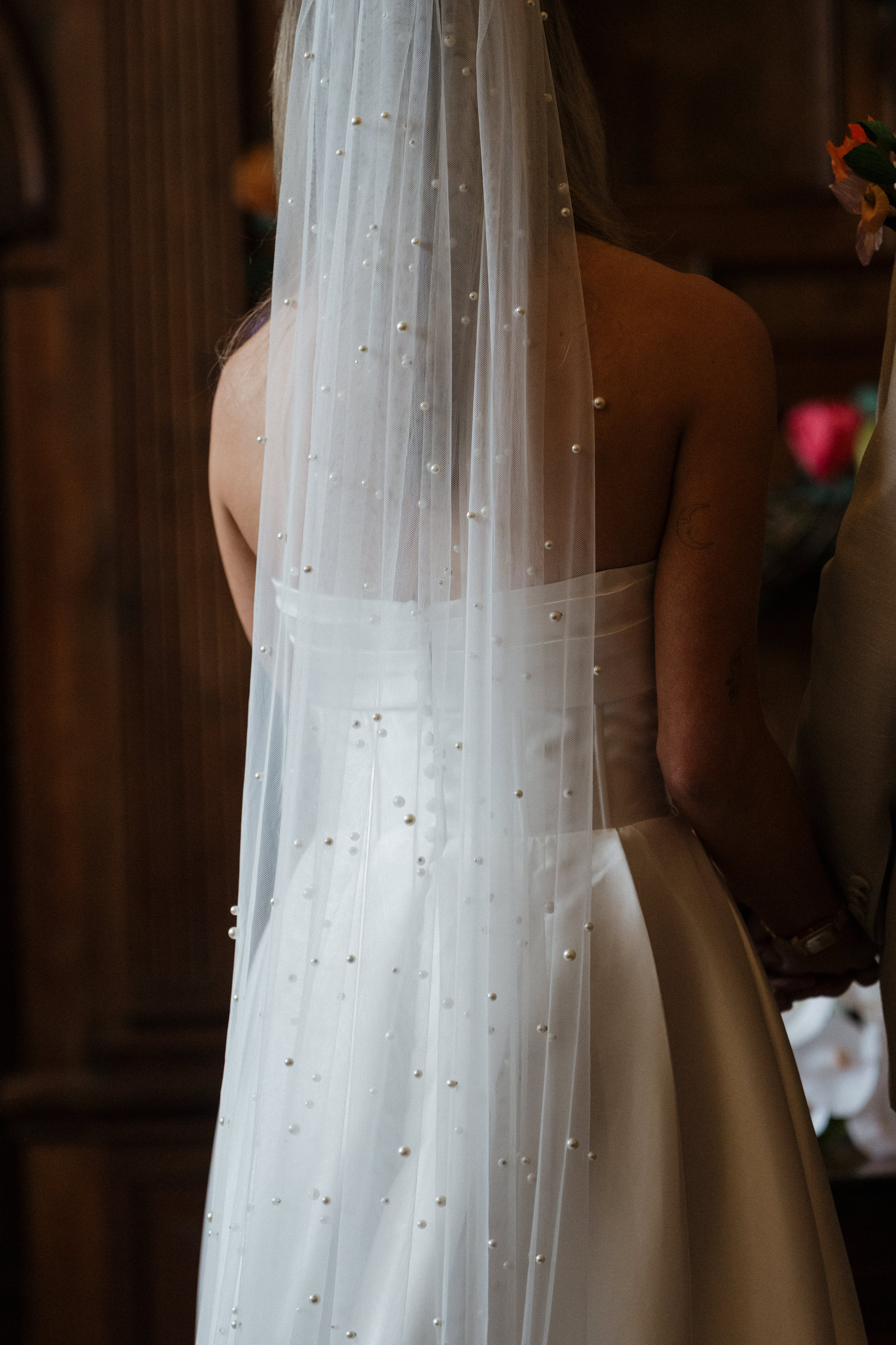 A bride wearing a strapless white dress and a veil with tiny pearls is holding hands with someone, both of them facing away from the camera.