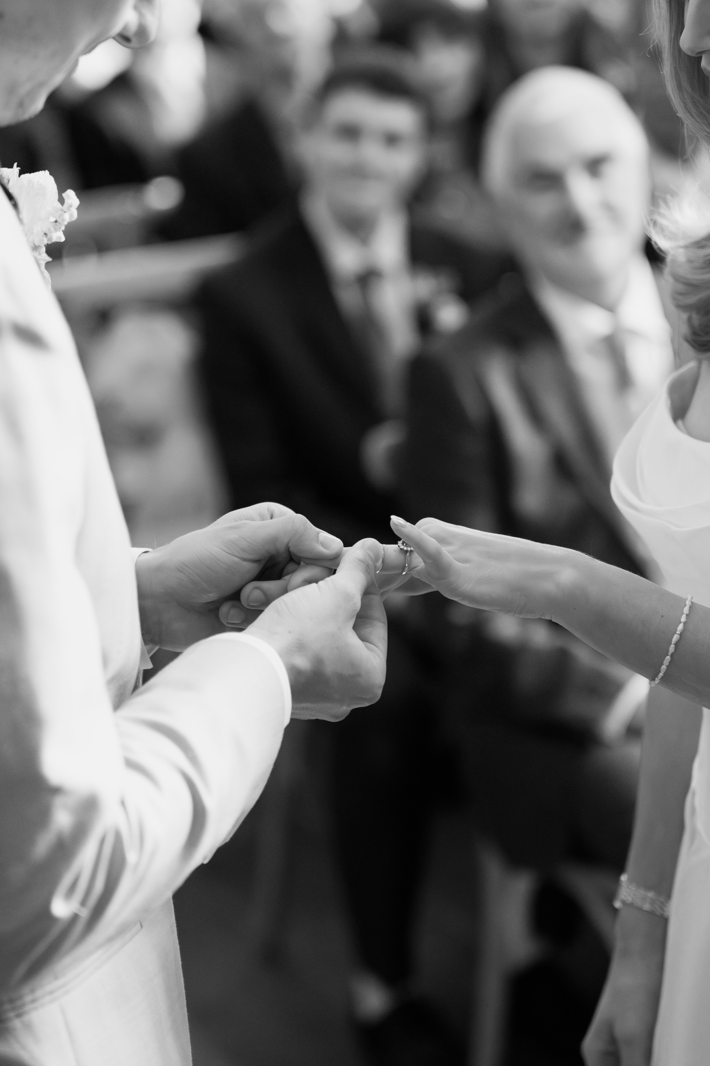 Someone is putting a ring on another person’s finger at a wedding, while guests sitting in the background appear out of focus.