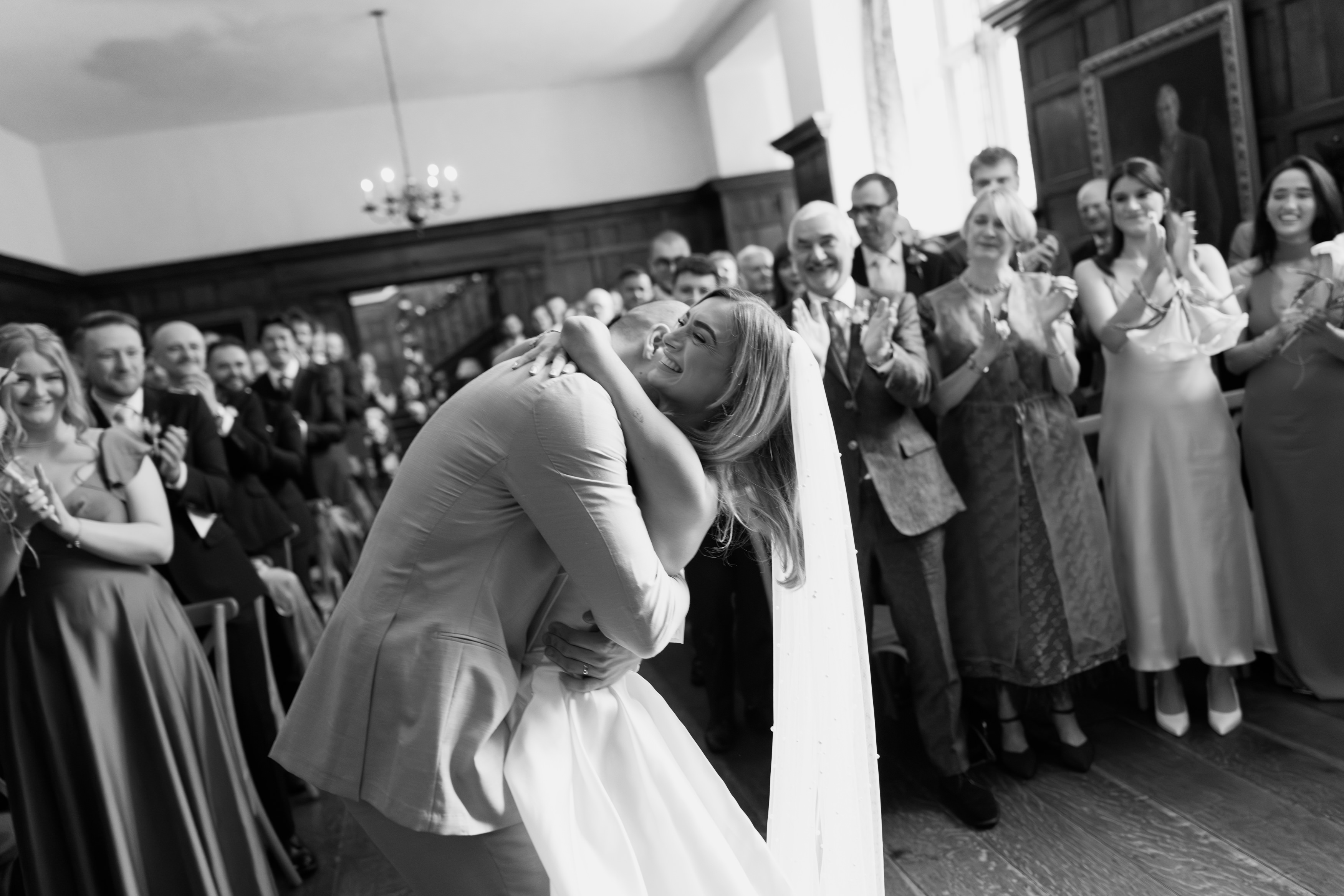The bride and groom hug and smile at their wedding as guests stand up and applaud in a room with wood-panelled walls.