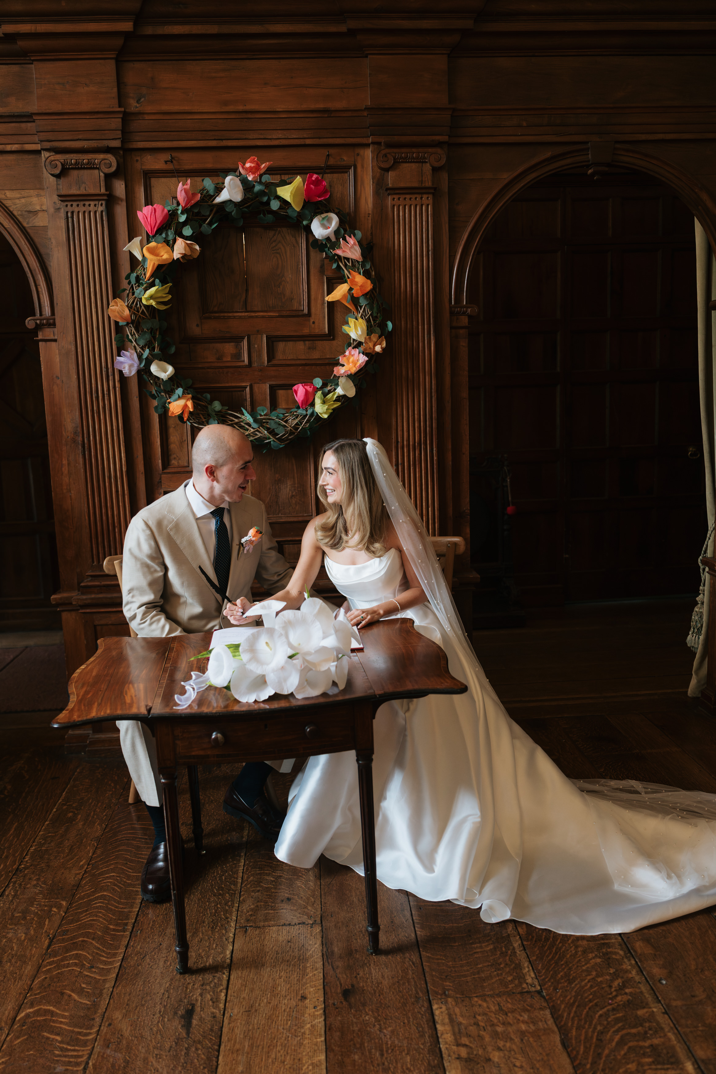 A bride and groom sit together at a wooden table in a fancy room with wood-panelled walls and a floral wreath, looking at each other.