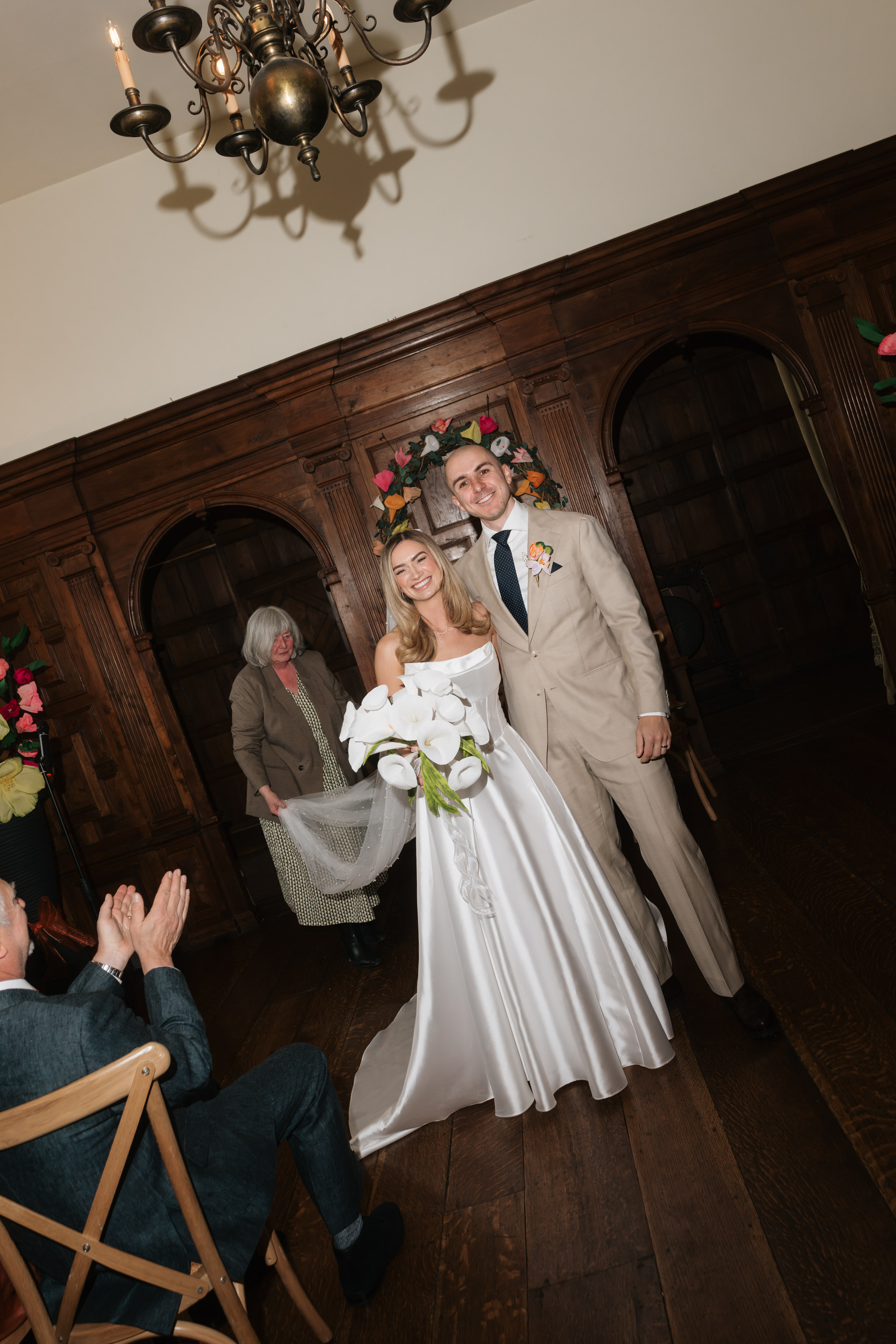 A bride in a white dress and a groom in a beige suit smile while standing indoors, holding a bouquet. A guest is applauding nearby, and someone else is walking behind them.