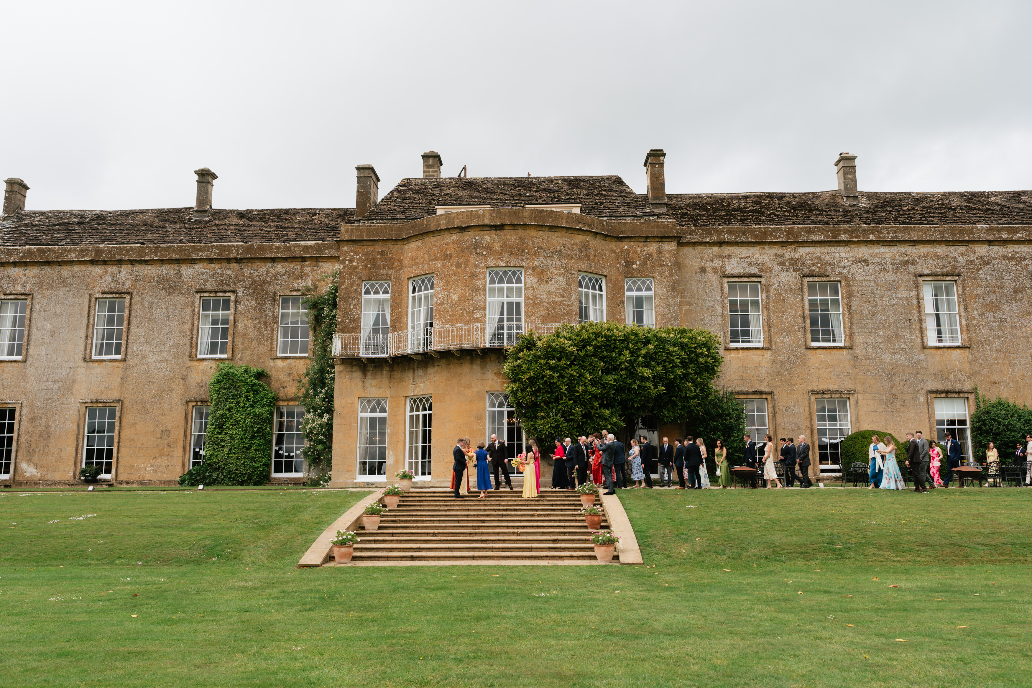 Some people are hanging about on the grass in front of a large, old stone building that has tall windows and a staircase in the middle.