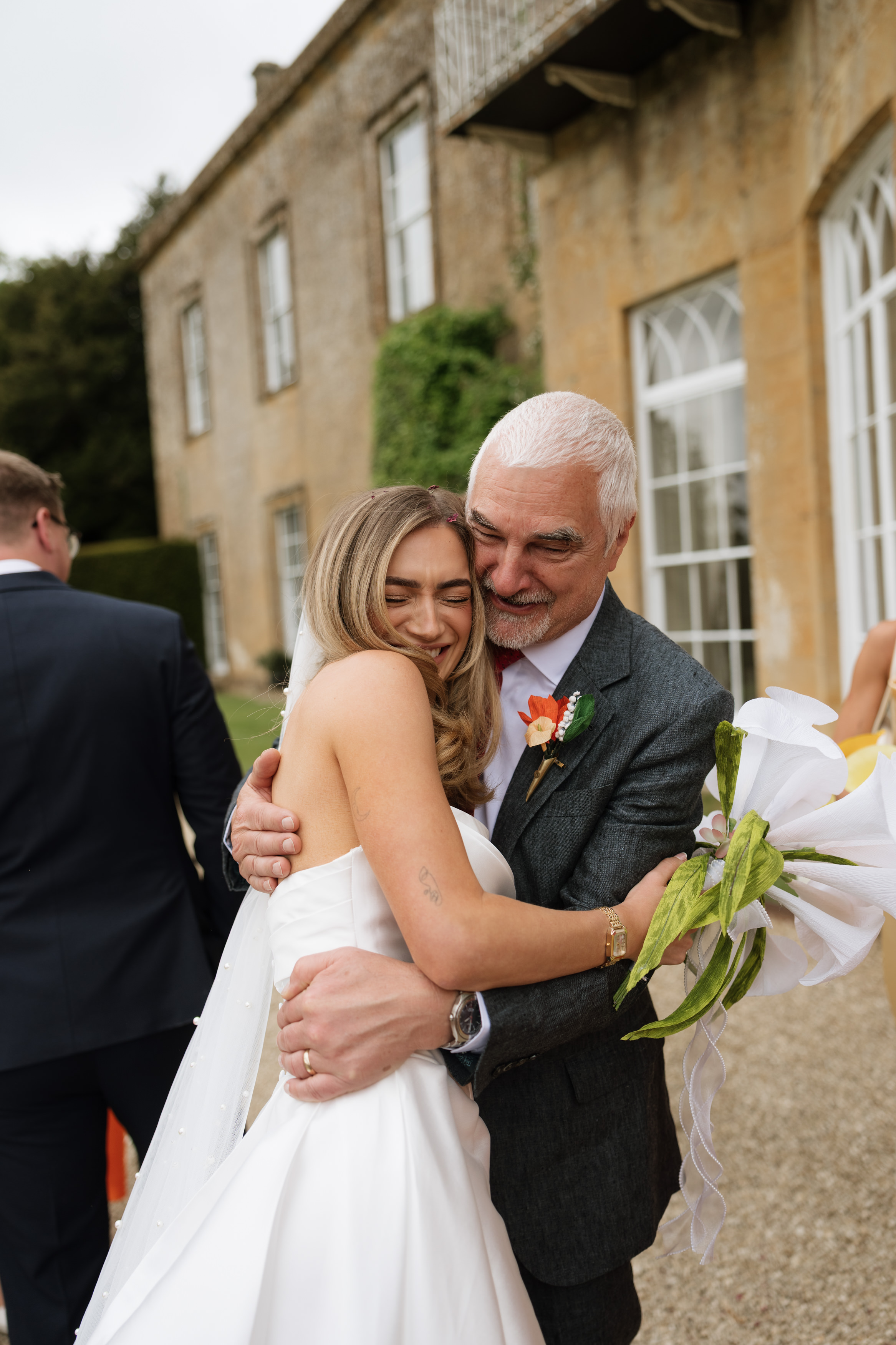 A bride wearing a white dress and veil hugs an older man in a suit outside a stone building, and they’re both smiling.