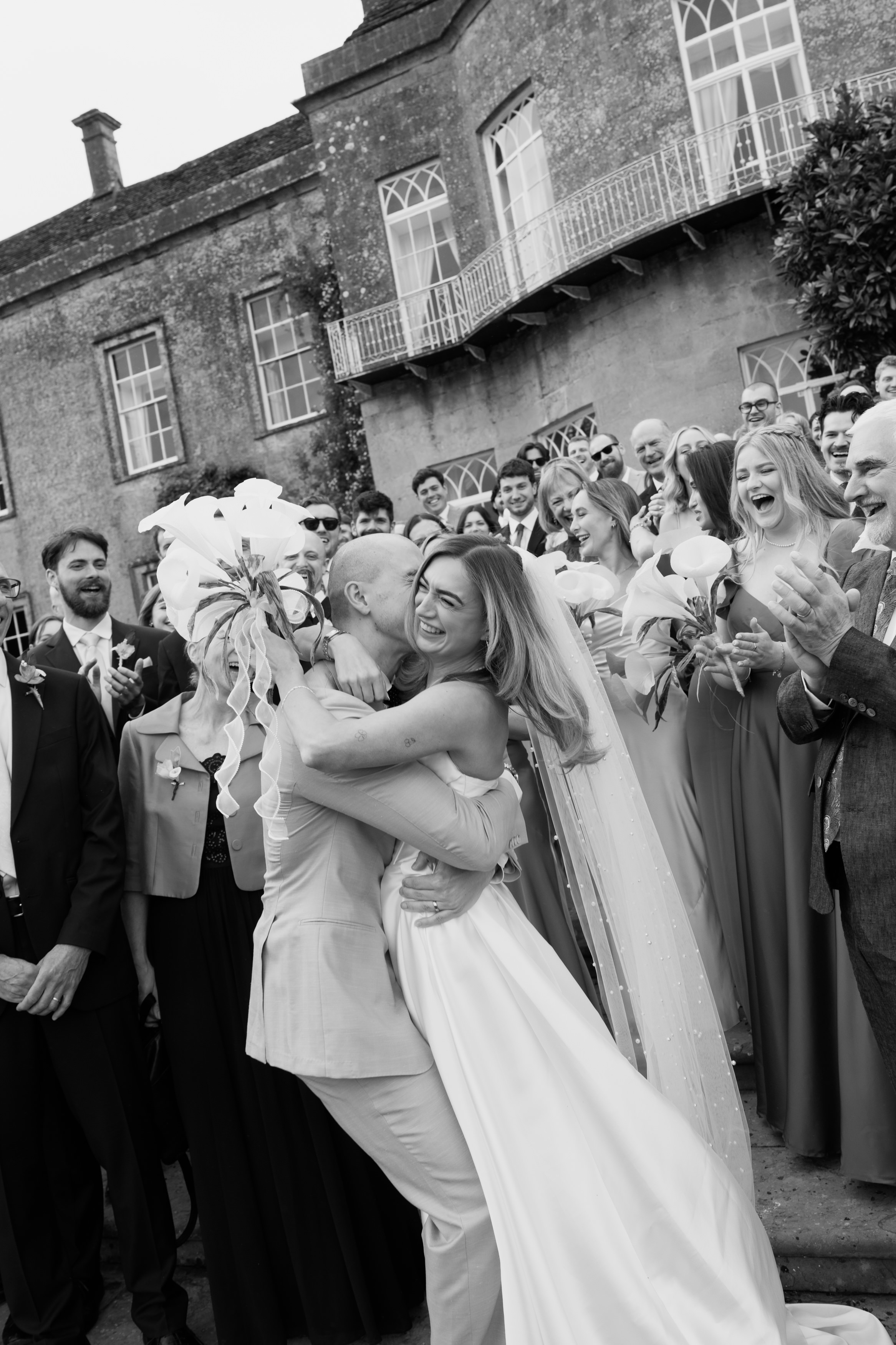 A bride and groom hug and smile outside whilst their friends and family, all dressed up, cheer for them in front of an old building.