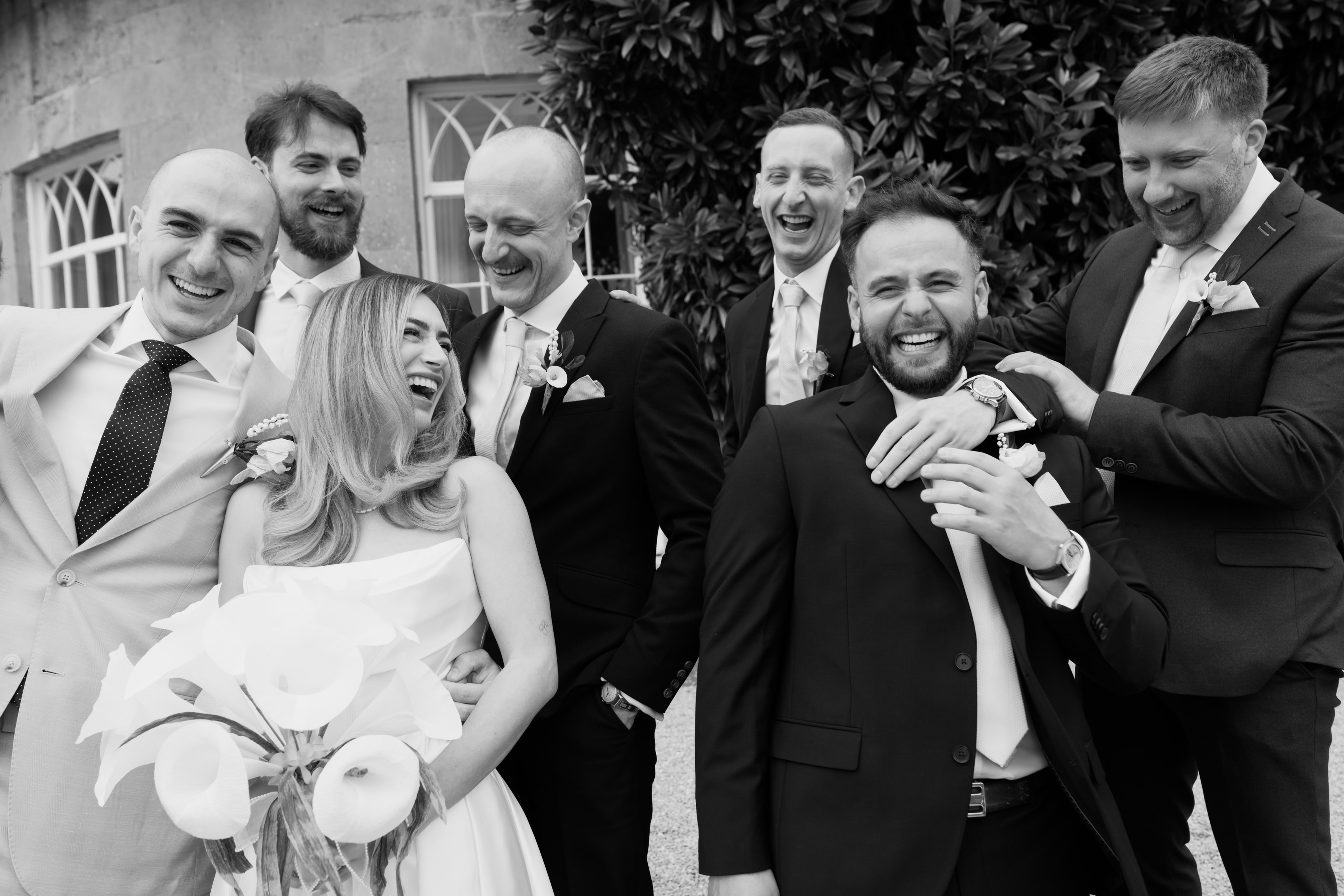 A bride holding calla lilies is laughing with five men in suits, all of them smiling and having a good time together outside near a building and some plants.